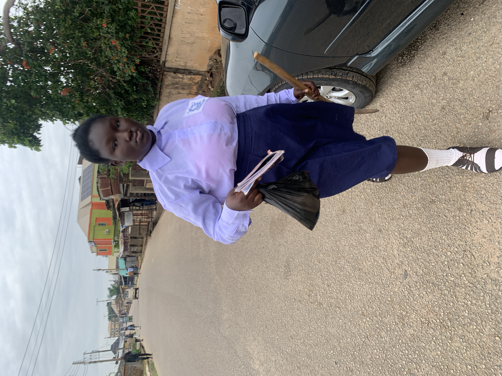 A young woman in a school uniform, consisting of a light purple button-down shirt and a dark blue pleated skirt, walks down a paved street in Jos, Nigeria. She carries a black plastic bag in her left hand, which appears to contain papers and possibly other items. In her right hand, she holds a light brown stick. She is wearing white socks and black and white shoes. A dark-colored car is parked to her right. The background shows a street scene with buildings, power lines, and other people walking in the distance. The sky is overcast.