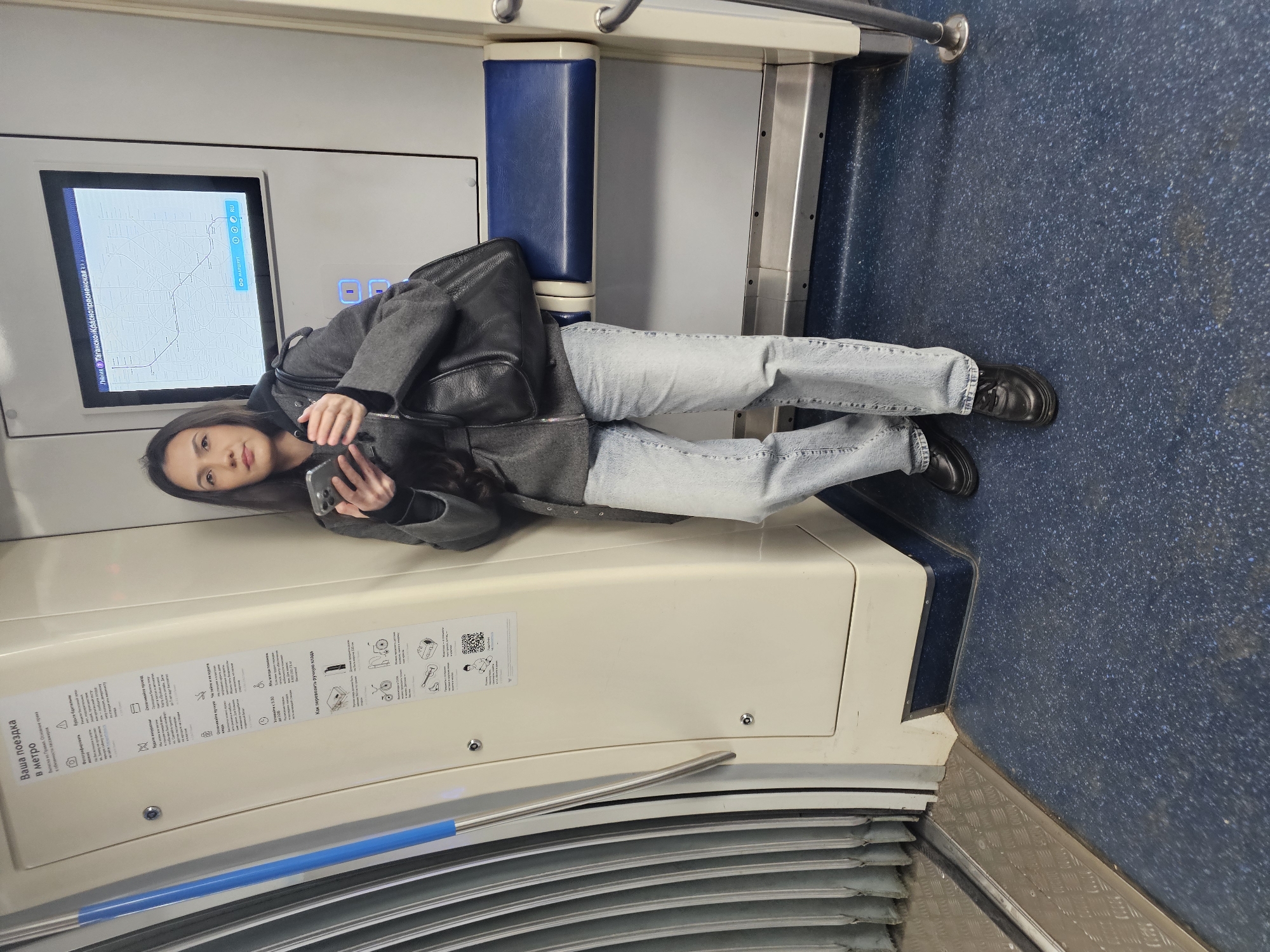The image shows a woman lying on a bench in what appears to be a subway car, likely in Moscow, Russia, based on the inscription "Лийния Таганско-Краснопресненская в МАРШРУТ RU" which translates to "Tagansko-Krasnopresnenskaya Line IN ROUTE RU". The woman is wearing jeans, a dark jacket, and dark shoes. She is holding a phone. The bench has a blue cushion and is situated next to a wall with a digital display showing a map, likely of the subway system. There are informational signs on the wall with text and pictograms. The floor has a speckled blue pattern. The overall environment seems clean and well-maintained.