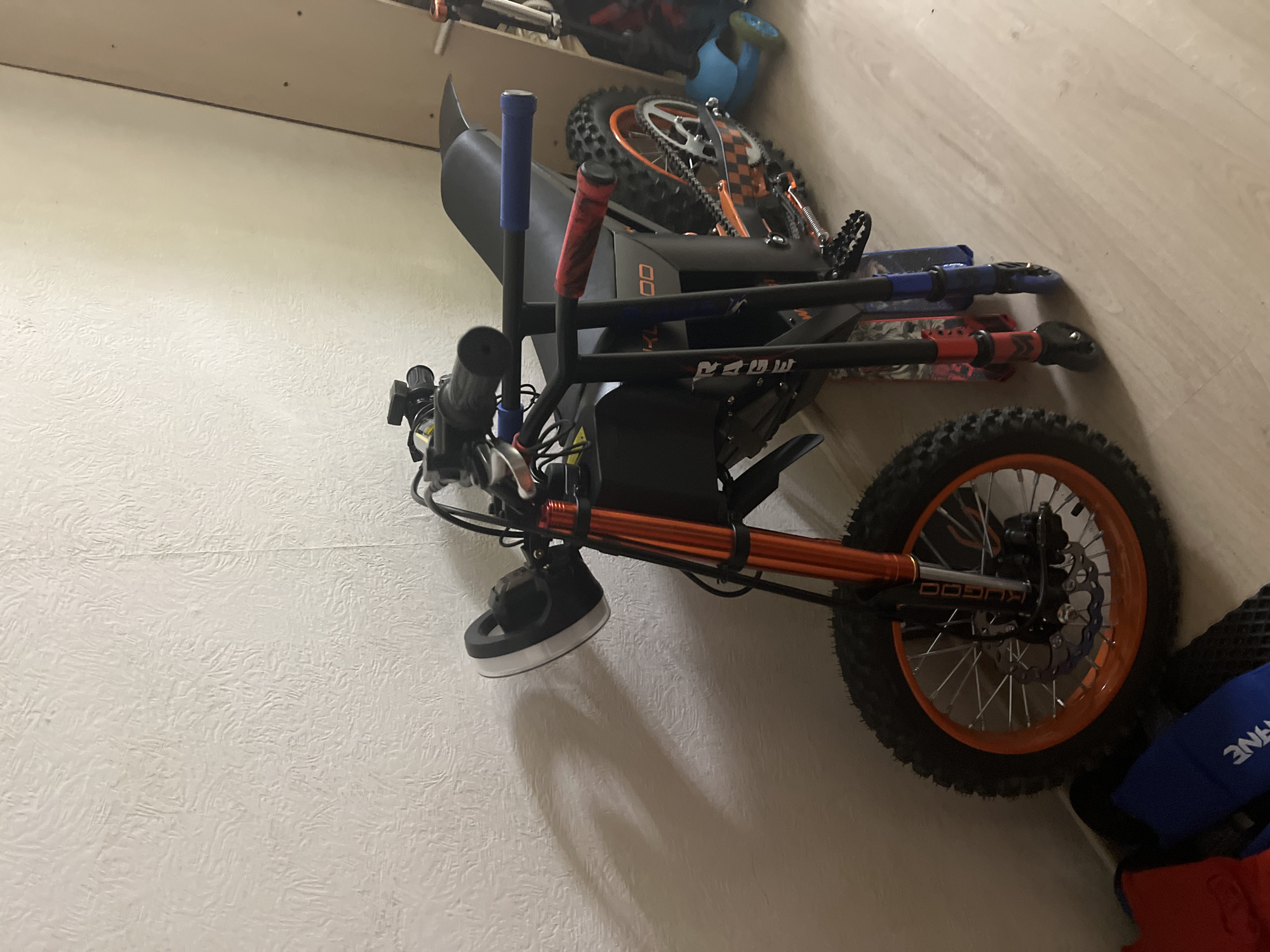 The scene depicts an indoor area featuring an electric dirt bike and two kick scooters, positioned against a textured off-white wall and a light-toned wooden floor. The primary object is a black electric dirt bike, characterized by orange front suspension forks, orange wheel rims, and an orange chain. The brand "KUGOO" is visible on the front fork. It features a round, black headlight with a white outer ring, a black seat, and aggressive knobby tires. A disc brake is present on the front wheel. To the right of the dirt bike, two kick scooters stand upright. One scooter has red handlebars and lower pole accents, while the other has blue handlebars and accents. The word "RAGE" is partially visible vertically on the black handlebar support of one scooter. In the bottom-left corner, a red fabric item and a blue item with white lettering (partially visible, possibly "CRANE" or similar) are on the floor. A vertical, light-brown panel or partition is visible in the background on the right. Illumination suggests a light source from the right, casting distinct shadows of the objects onto the wall to the left. No individuals are present in the image.