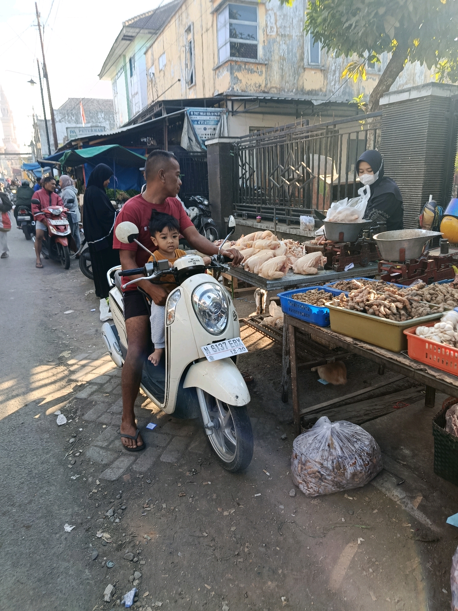 In Gondanglegi, Indonesia, a man in a red t-shirt and dark shorts rides a cream-colored Honda scooter, holding a young child in his lap. The child wears a mustard-colored shirt and grey pants. The scooter's license plate reads "N 6137 EBX". They are on a street lined with market stalls.
To the right of the scooter, a market stall displays whole raw chickens on a table. Behind the chickens, a woman wearing a black hijab and a white face mask is seen interacting with a scale. Further to the right, another table holds various produce, including what appears to be ginger and garlic in baskets. A large plastic bag filled with what looks like dried produce or debris sits on the ground.
In the background, other market-goers and vendors are visible, some on motorbikes, and buildings with worn facades line the street under a bright, sunny sky. Shadows from trees and structures are cast across the ground, indicating it's likely late morning or early afternoon. The overall scene depicts a typical day at a local market in Indonesia.