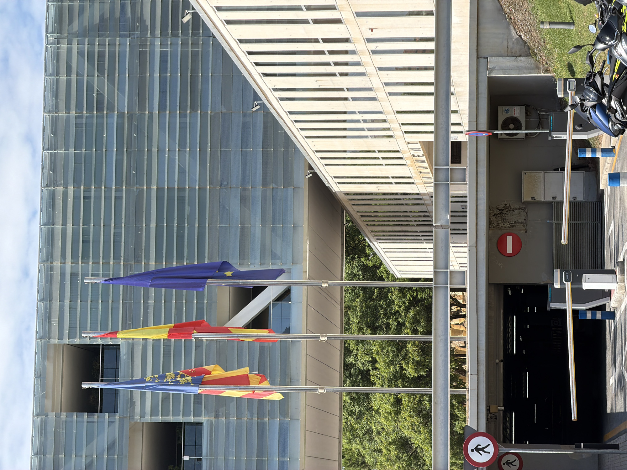 The image shows the entrance to a parking garage with three flags flying in front of a modern building. The flags, from left to right, appear to be the flag of the Valencian Community, the flag of Spain, and the flag of the European Union. The building behind the flags is a multi-story structure with a facade of horizontal glass panels. To the right of this building, another section of the building is visible, constructed with light-colored concrete and featuring numerous vertical windows. In front of the parking garage entrance, there are two red circular signs, one with a pedestrian symbol and another with a horizontal red bar, indicating restrictions. A yellow and white barrier arm is lowered, blocking entry to the parking garage. Several security cameras are mounted on the buildings. At the bottom right of the image, two motorcycles are parked. The image is taken on a bright, sunny day with a partly cloudy sky. The text "AYUNTAMIENTO" (City Hall) is visible on the concrete building, suggesting the location is a municipal building. Based on the presence of the Valencian Community flag, the location is likely in the Valencian Community region of Spain, and specifically, the architecture and context suggest it could be the City Hall of Benidorm, Spain.