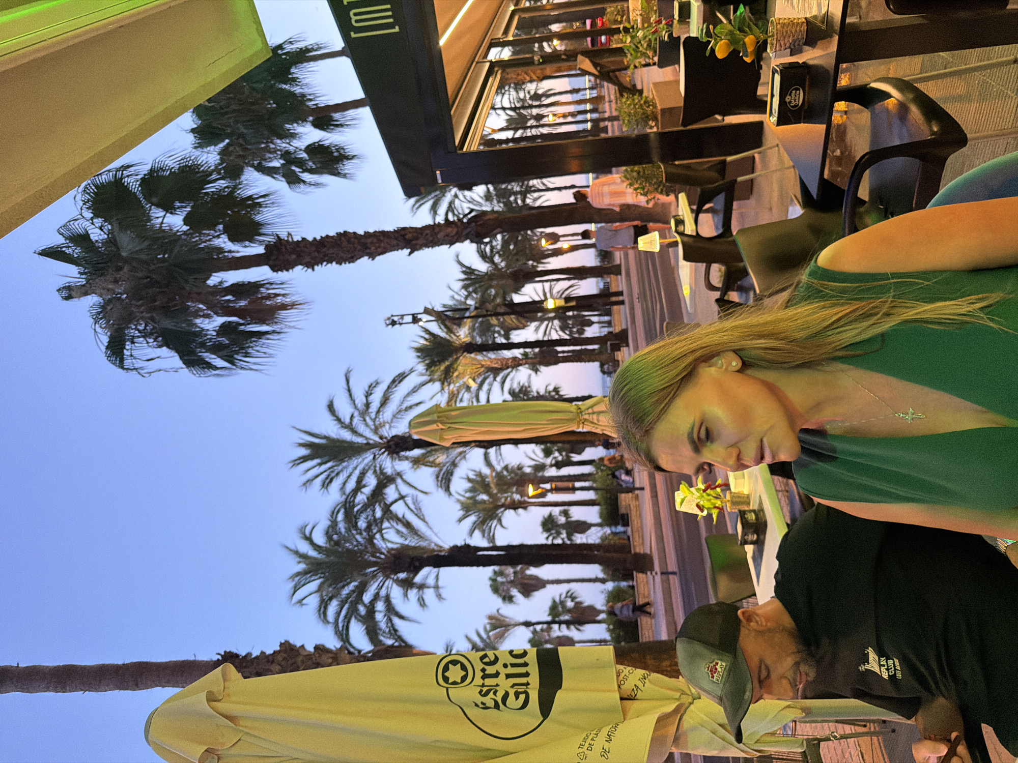 The image depicts a scene at an outdoor dining establishment in Salou, Spain, likely near the waterfront, given the presence of palm trees and a promenade. The time of day appears to be dusk, with a soft twilight sky. In the foreground, a woman with blonde hair and a green dress is seated at a table, looking to her left. To her left, a man wearing a black t-shirt and a baseball cap is also seated at a table, looking down at his phone. Both are at a cafe or restaurant with outdoor seating. Several tables and chairs are visible, some unoccupied. Palm trees line the promenade behind the seated couple, and a building with an awning, possibly part of the establishment, is visible on the right. A street with a few people walking and cars passing is in the background. The atmosphere suggests a relaxed evening at a popular tourist destination.