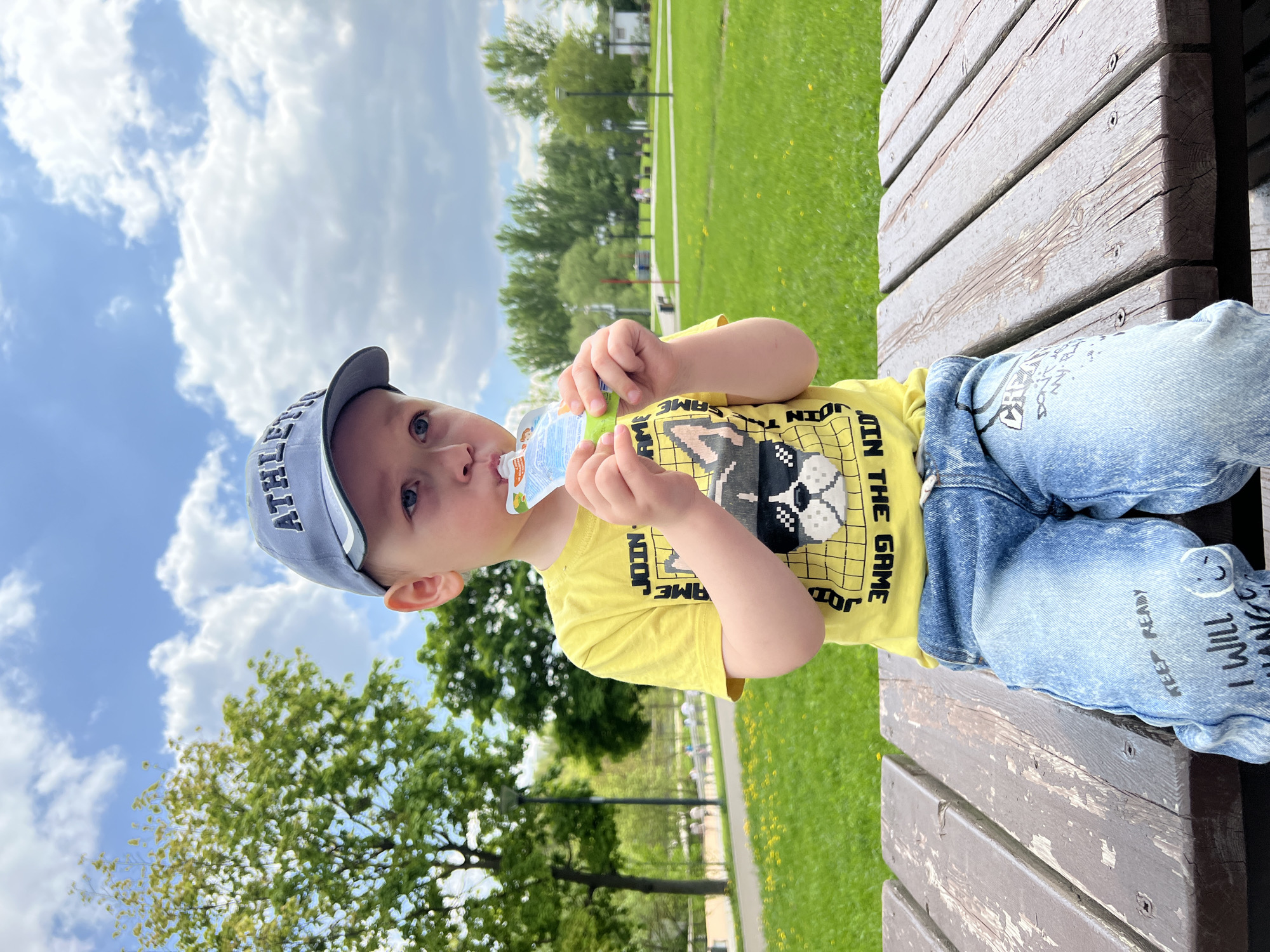 A young child in a grey "Athletic" baseball cap, yellow shirt with a dog graphic, and blue jeans sits on a wooden park bench, drinking from a clear plastic bottle with a green cap. The child is facing the camera. The background consists of a grassy park area with trees and a walkway. The sky is visible, with blue patches and fluffy white clouds. The location is potentially in a park in the city of Moscow, Russia.