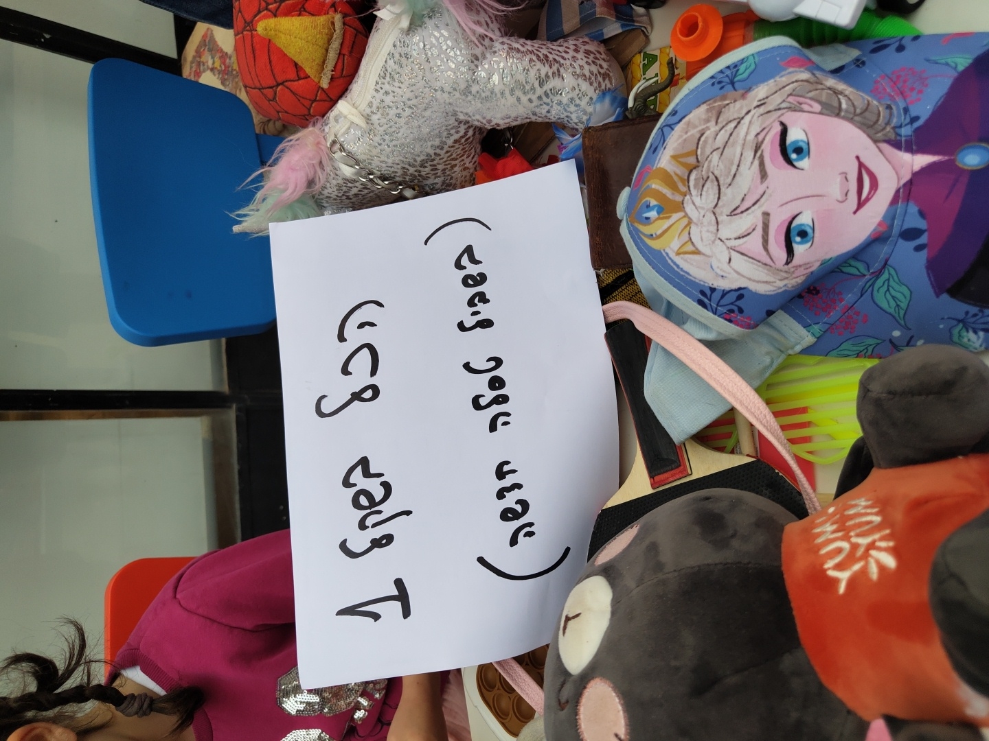 A close-up, slightly angled, indoor shot shows a collection of toys and objects with a child partially visible in the lower left corner. The child is wearing a magenta-colored shirt with silver heart sequins.
Dominating the center of the frame is a white sheet of paper with Hebrew writing, presumably a sign or message. The toys and objects surround the paper. These include a blue child-sized chair, a plush Spiderman head, a silver unicorn plush, a cap with an image of Elsa from Frozen, a plush black animal with pink details, and other miscellaneous items.
The background is slightly blurred, suggesting an indoor setting. There is no visual information to confirm that the location is Tel-Aviv, Israel.