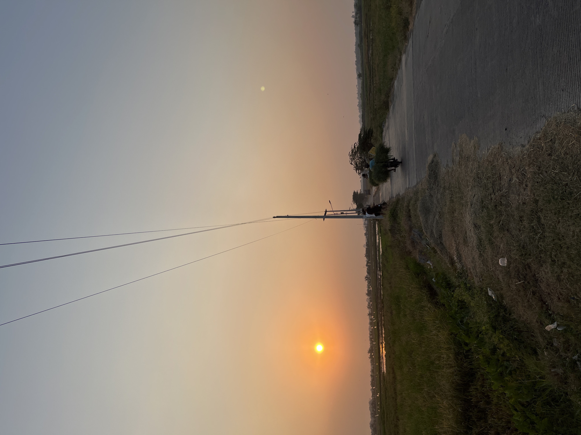 A wide shot captures a rural scene at sunset in Pecangaan, Indonesia. The sun, a bright orange orb, is low in the sky to the left, casting a warm glow over the landscape. Power lines stretch diagonally across the upper portion of the frame. A paved road runs from the foreground towards the middle ground, where a person on a motorcycle is transporting a large bundle of green foliage. To the left of the road, a field of green grass slopes down towards a body of water, likely a rice paddy, with scattered white markers. In the background, a line of trees and some structures are visible. A utility pole stands beside the road, with streetlights and smaller wires attached. Several other figures are present near the utility pole. The ground along the side of the road is covered in dry grass and some scattered white plastic debris. The sky is a gradient of soft orange and pale blue.