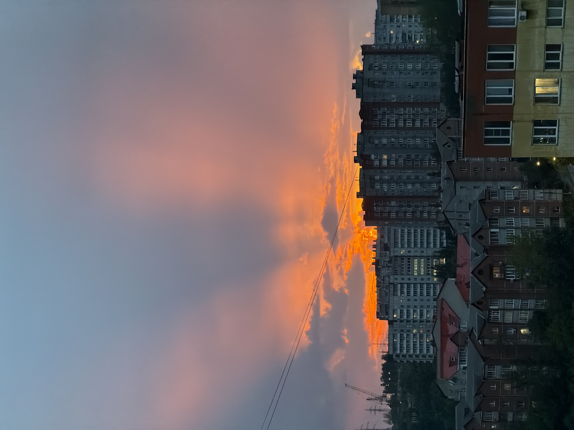 The image depicts a cityscape at dusk, likely in Krasnoyarsk, Russia, given the architectural style of the buildings and the presence of power lines. The sky is dramatically lit by a vibrant sunset, with hues of orange and pink radiating from behind the clouds. Multiple high-rise apartment buildings, typical of Soviet-era urban planning, dominate the middle and background. Some buildings are illuminated with interior lights, suggesting that it is evening. In the foreground, there are several lower-rise residential buildings and trees, casting shadows. Power lines are visible spanning across the sky, and industrial structures, including a crane and power pylons, are discernible in the distance on the left. The overall scene conveys a sense of urban life at the end of the day, with the dramatic natural light providing a striking contrast to the man-made structures.