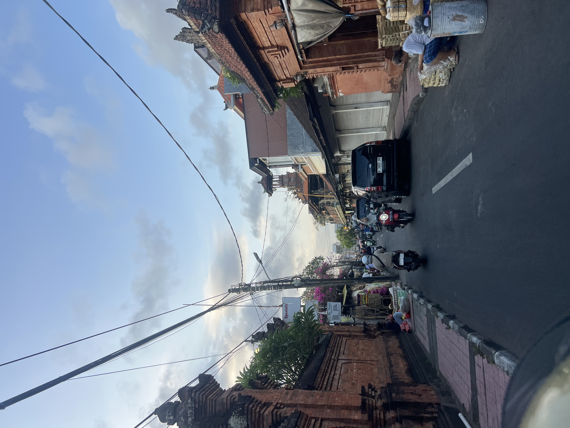 The image depicts a street scene in Denpasar, Indonesia. A narrow road is lined with traditional Balinese architecture, characterized by ornate brick walls and tiled roofs. Power lines crisscross the sky above. On the left side, a brick wall borders the street, with lush green foliage growing over it. Further down the street, there are various shops with closed roll-up doors and some open stalls selling goods. Several people are present, with two individuals sitting on the sidewalk on the left, and a person sitting on the right next to a collection of baskets and a metal bin. The street is active with traffic, primarily consisting of motorbikes and a black SUV. People on motorbikes are moving along the road, some wearing helmets. The sky above is partly cloudy, with a soft, diffused light suggesting it might be early morning or late afternoon. The overall atmosphere is that of a bustling street in a Southeast Asian city.