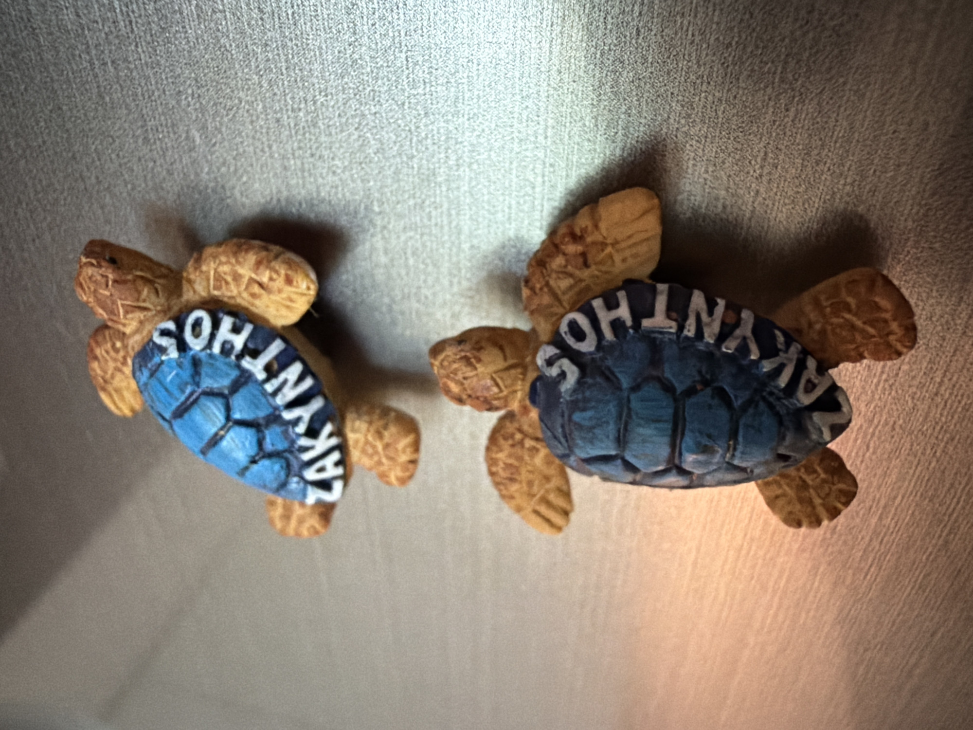 Two small, decorative sea turtle figurines are positioned on a textured, metallic surface. The turtles are made of a brown, textured material resembling a shell, with blue shells that have white lettering. The upper turtle's shell reads "ZAKYNTHOS" in white capital letters, and the lower turtle's shell also appears to have "ZAKYNTHOS" written on it, though it is partially obscured and harder to read. Both turtles are oriented with their heads facing away from the viewer and their flippers extended. The lighting in the scene creates a soft gradient on the surface, with a brighter, warmer tone on the left and a darker, cooler tone on the right.