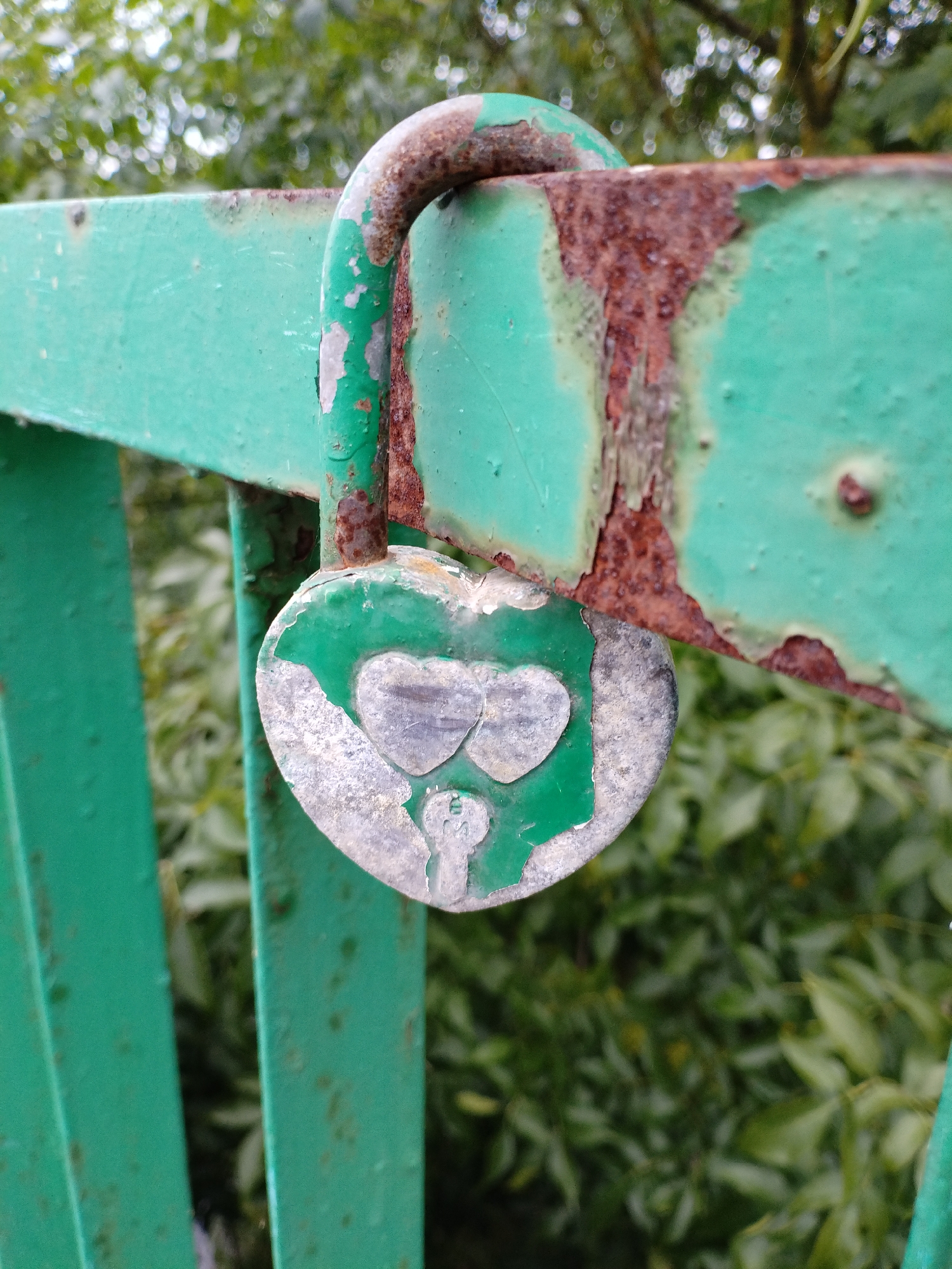 A heart-shaped padlock with green paint chipping off, revealing a silver surface, is attached to a rusted green metal railing. The railing is part of a structure, likely a bridge or fence, with visible signs of rust and wear. Green foliage is visible in the background, out of focus. The image is a close-up shot, focusing on the lock and a section of the railing. This scene is typical of a public space where locks are sometimes placed as symbols, potentially on a bridge in the city of Bialynichy, Belarus.