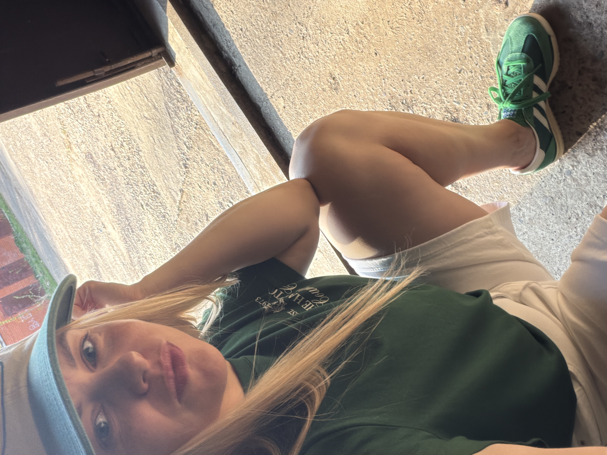 A selfie-style photograph shows a young woman lying on an outdoor concrete surface. She is wearing a light blue baseball cap, a dark green t-shirt with a printed design, and light-colored shorts. Her legs are crossed, and she is wearing a green and white Adidas sneaker on her right foot. The background includes a section of weathered concrete pavement and a dark, undefined structure overhead. There is insufficient data to provide the location in Votkinsk, Russia.