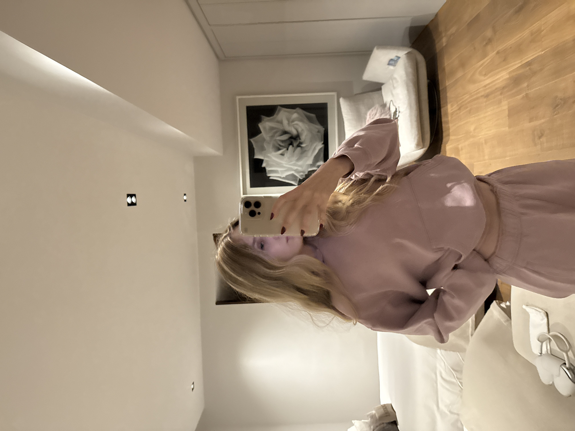 A young woman takes a selfie in a bedroom. She is wearing a pink outfit and holding a cell phone. The background shows a framed black and white rose photograph, a white sofa, and a wooden floor. The location is likely a hotel room or apartment in Mexico City, Mexico, based on the modern interior design and the presence of a Mexican power outlet visible near the bed. The woman appears to be in the process of taking a photo of herself in the mirror.