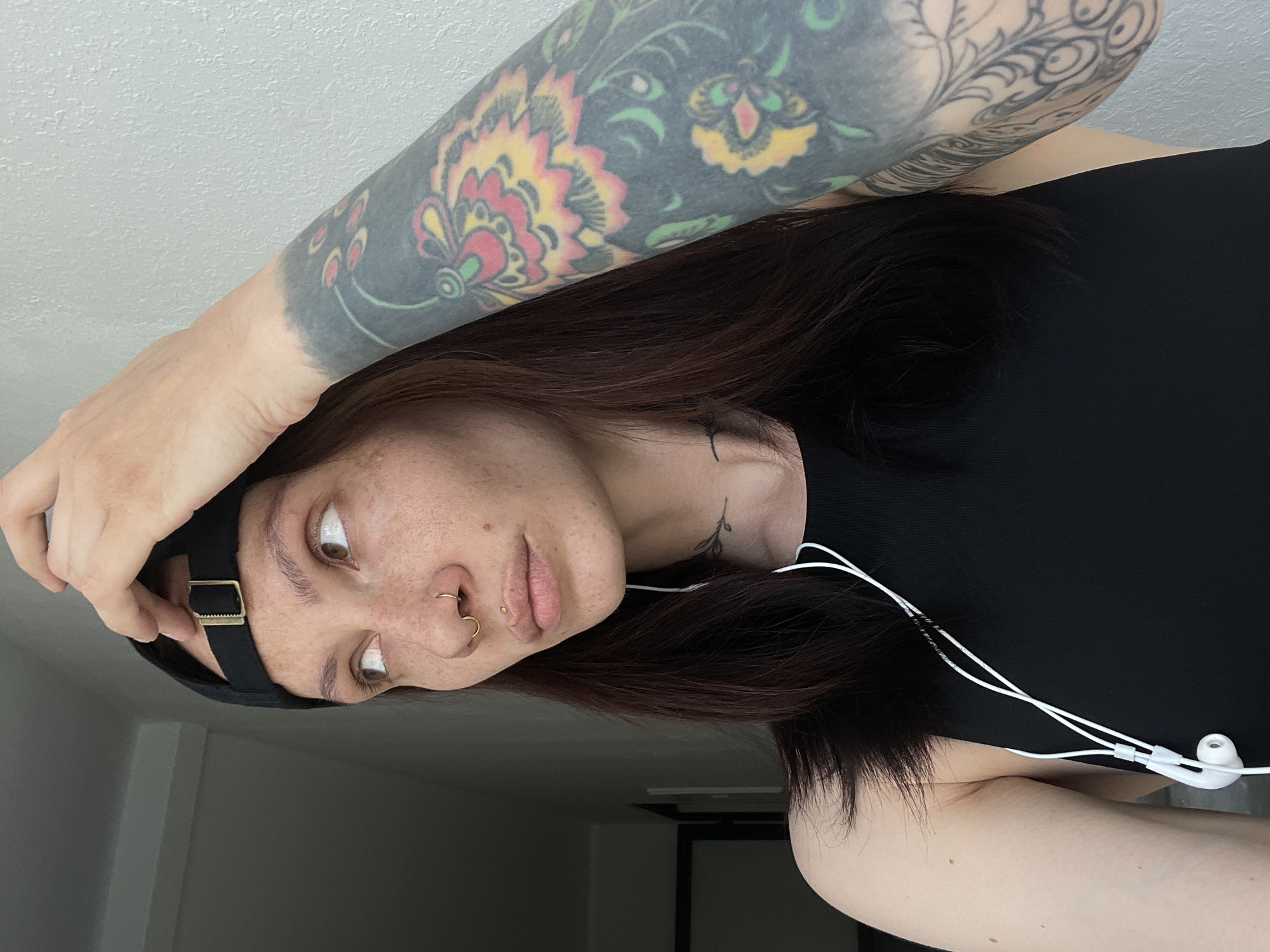 A fair-skinned person with brown hair is shown indoors, wearing a black tank top, a black cap with a buckle detail, and white earphones. The person has a tattoo on their right arm. They have multiple piercings on their nose. Another tattoo is visible on their neck. The background is a white wall and a blurred view of a door and an unknown room. Location information not available.