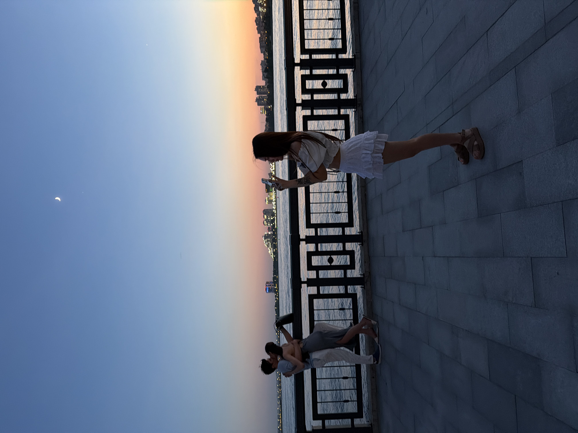 The image shows a woman in a white outfit looking at her phone while standing by a metal railing with a city skyline in the background. Below, a couple embraces near the railing. The sky transitions from blue to a sunset hue. The setting appears to be a waterfront area.