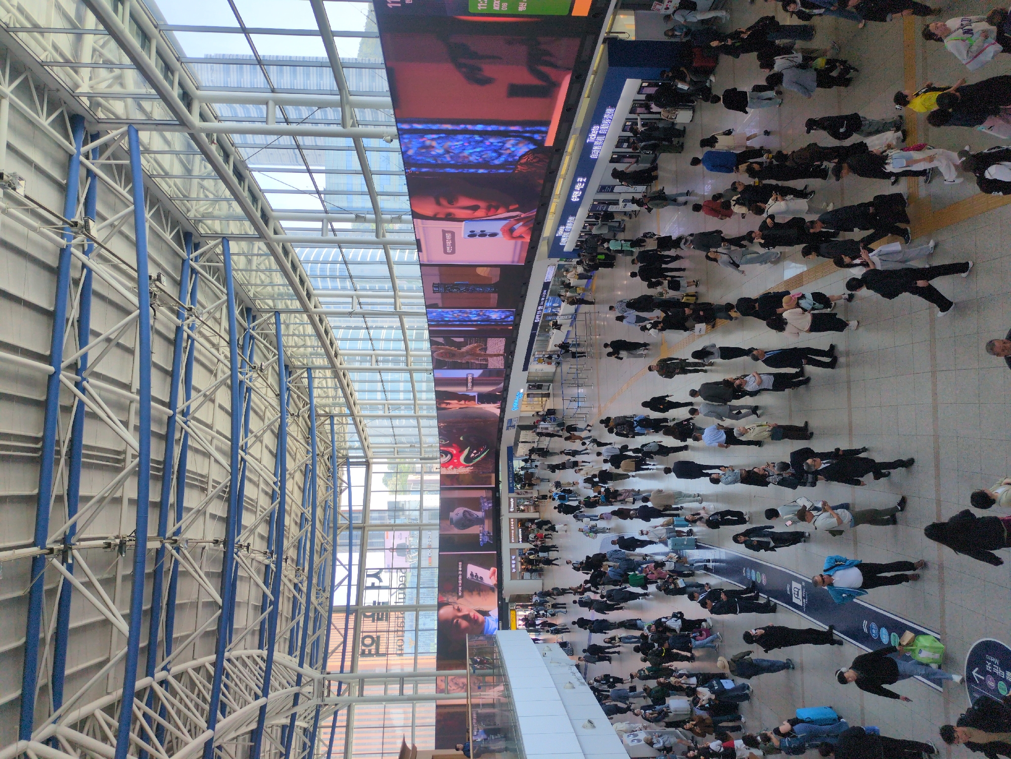 This image shows the interior of a busy transportation hub in Seoul, South Korea. A large crowd of people is seen on a tiled floor. Overhead, a high ceiling with a blue and white structural framework and transparent panels is visible. A large LED screen displays advertisements and digital content. The scene suggests a bustling environment with people likely commuting or traveling. Signage in Korean characters is visible, along with the words "Metro" and "Tickets".