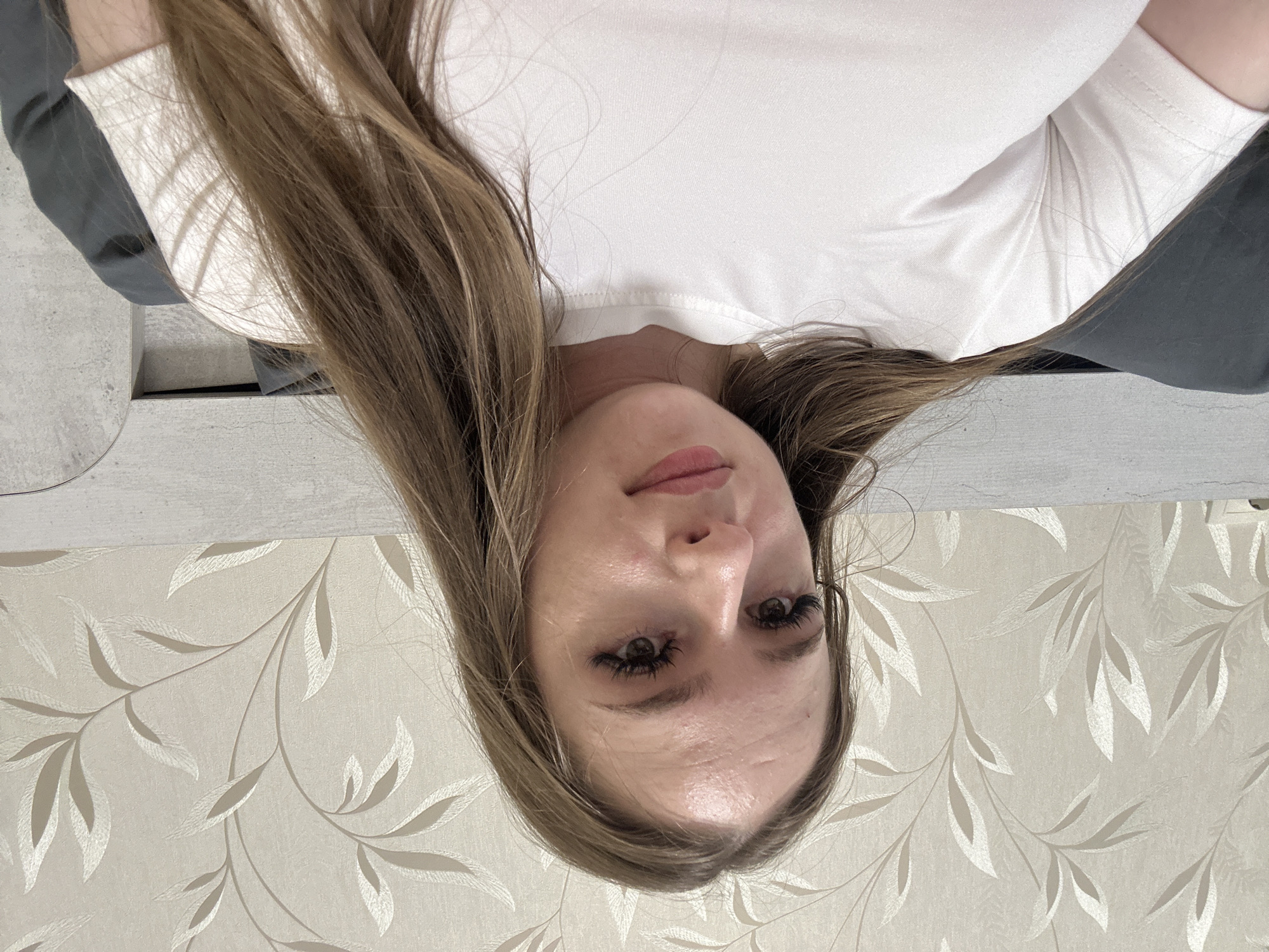 A young woman with long, light brown hair is shown in a close-up, frontal shot. She is wearing a white t-shirt. Her head is slightly tilted, and she is looking directly at the camera. She has on significant eye makeup, including false eyelashes. The background consists of wallpaper with a subtle floral pattern and a light-colored headboard. The lighting appears to be artificial and soft. The image is indoors, likely in a bedroom setting.