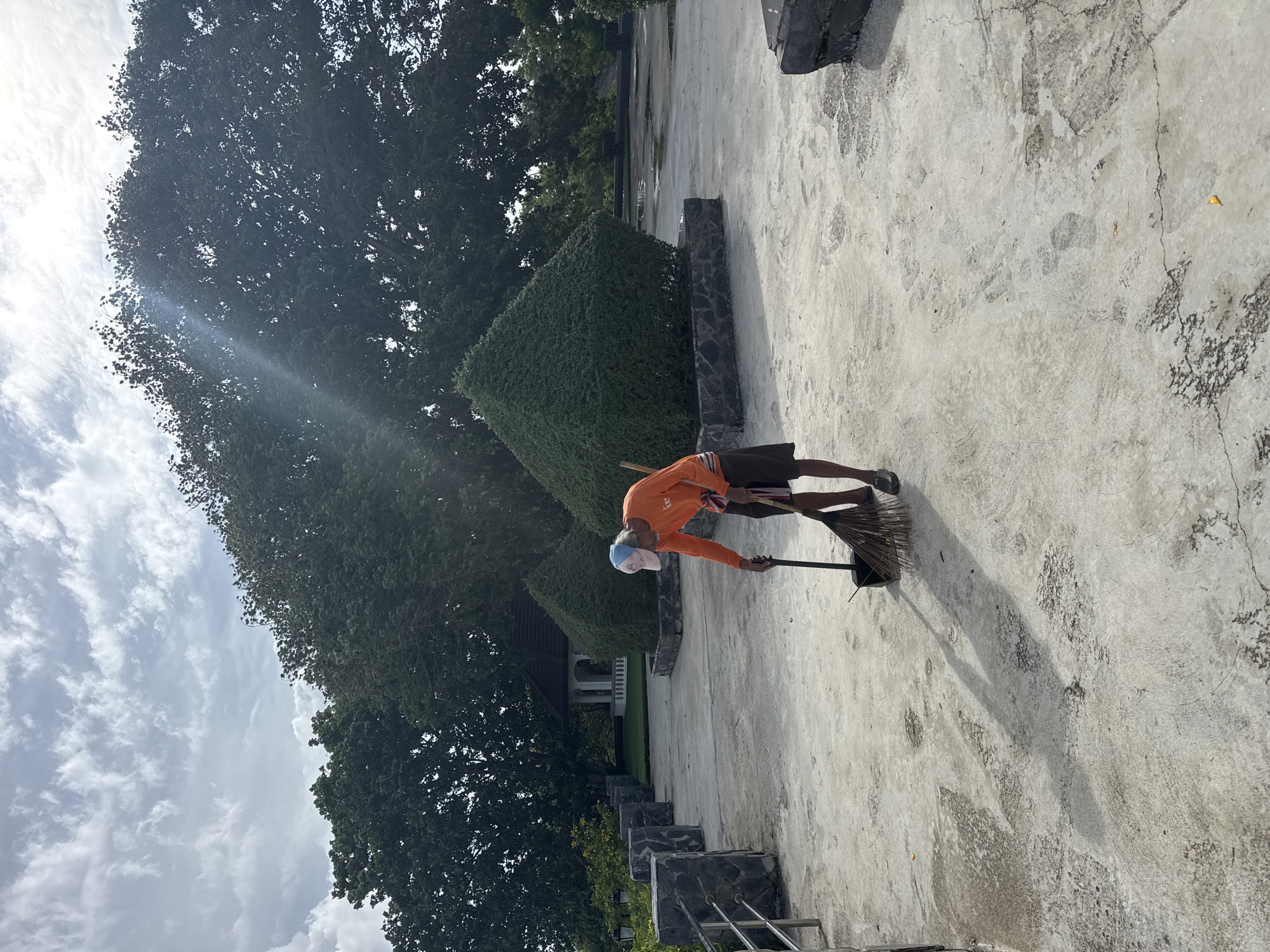 A man in an orange shirt and dark shorts is sweeping a concrete patio with a broom. He is wearing a blue and pink baseball cap. The patio is surrounded by stone walls and manicured bushes. In the background, there is a white gazebo and large trees. The scene is located in Rawai, Thailand. The sky is cloudy with sunlight breaking through.