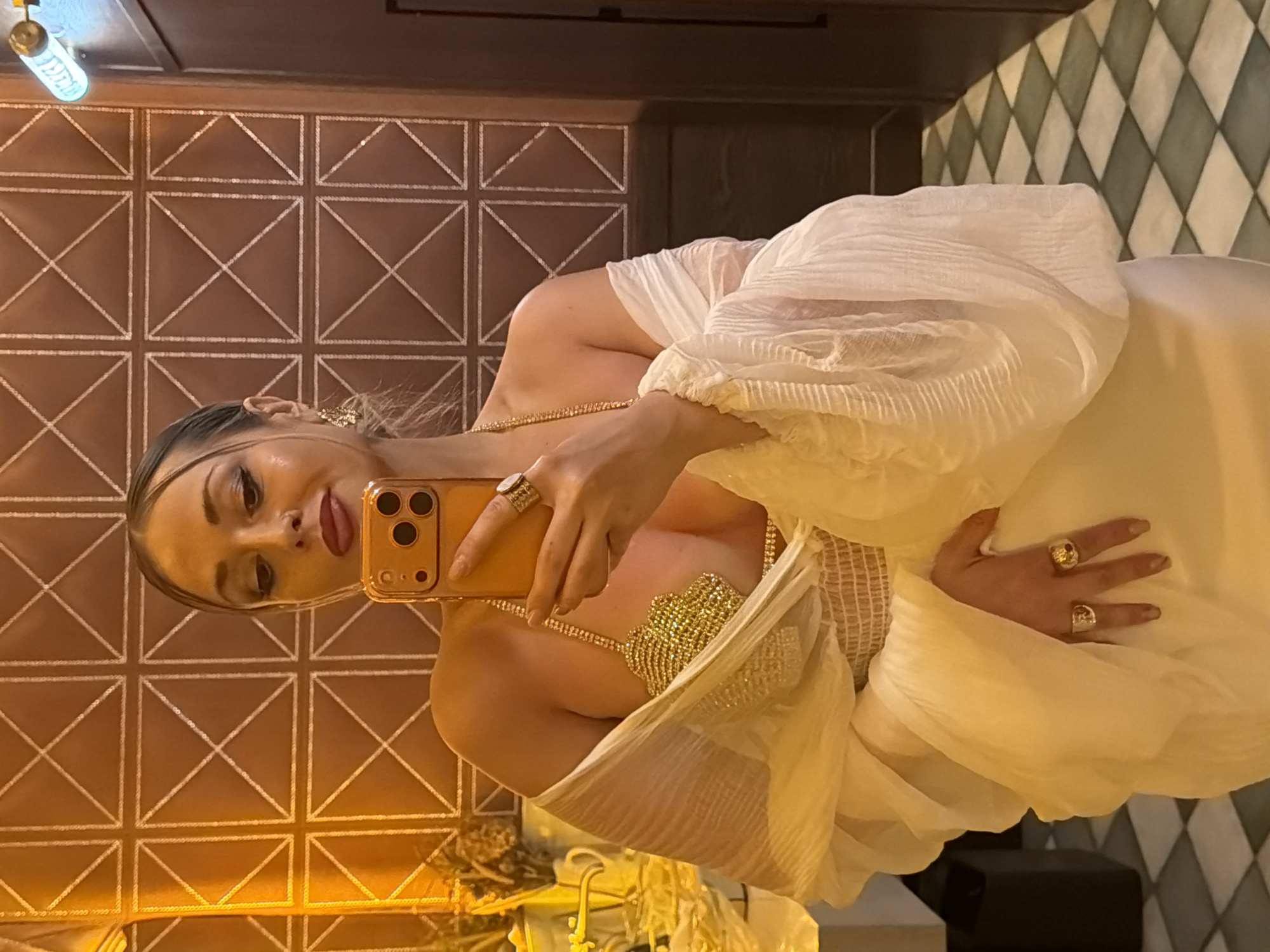 An indoor mirror selfie captures a woman holding an orange smartphone with a triple-lens camera system. The woman has light skin, dark eye makeup, and dark red lipstick, with her hair styled away from her face and strands framing her temples. She is wearing a revealing gold, jewel-encrusted bra top, and a loose, off-the-shoulder white or cream-colored garment with large, pleated sleeves. Her right hand, holding the phone, displays two large gold rings, and a third gold ring is visible on her left hand. She also wears gold dangle earrings and a delicate gold chain around her neck and torso. The background features a warm, brownish-orange wall with a raised white geometric grid pattern. The floor is tiled in a checkered pattern of white and dark green or black diamonds. A tubular light fixture emitting a blue glow is visible on the upper right side of the image, mounted on a dark brown wall panel. Blurred objects, possibly part of a countertop and decor, are visible on the far left. This scene is located in Krasnoyarsk, Russia.