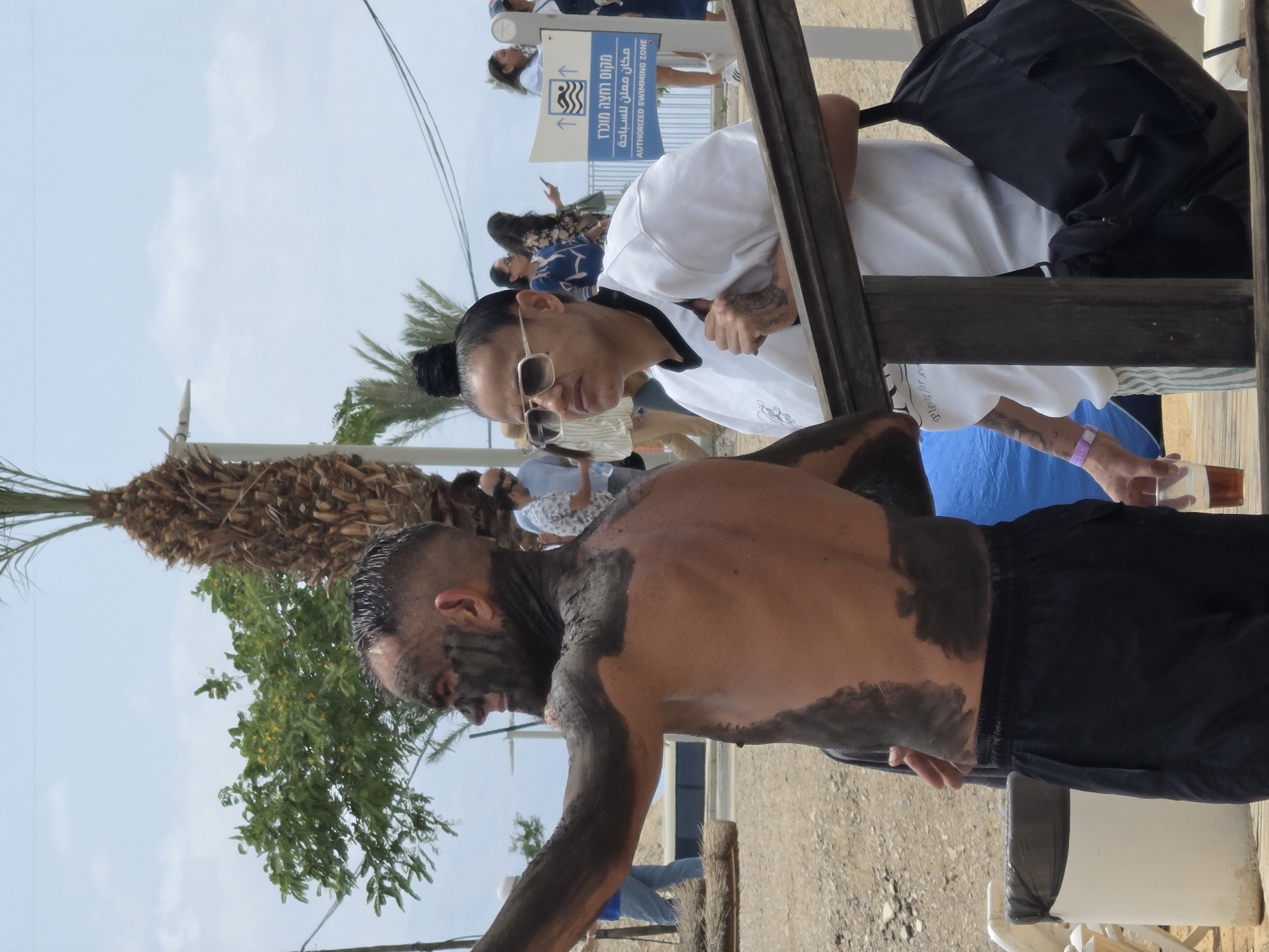 A horizontal eye-level outdoor shot shows a man partially covered in mud in Tamar Regional Council, Israel, with another person in the background and a sign visible.
In the foreground, a man with a shaved head and stubble stands facing left. His upper body and arms are covered in dark mud. He is wearing dark shorts. To the right, a chair is partially visible.
Behind him and slightly above, a person with dark hair pulled back in a bun wears sunglasses, a white shirt with a dark collar, and has tattoos on their forearms. They are looking down at the man with mud. A blue blanket or cloth is visible on their lap. A wooden structure is in the foreground.
In the background, other people are visible, along with a sign in Hebrew and English that says "Authorized Swimming Zone." A tree and a palm tree are on the left. The sky is blue with some clouds. The ground appears to be dirt or gravel.