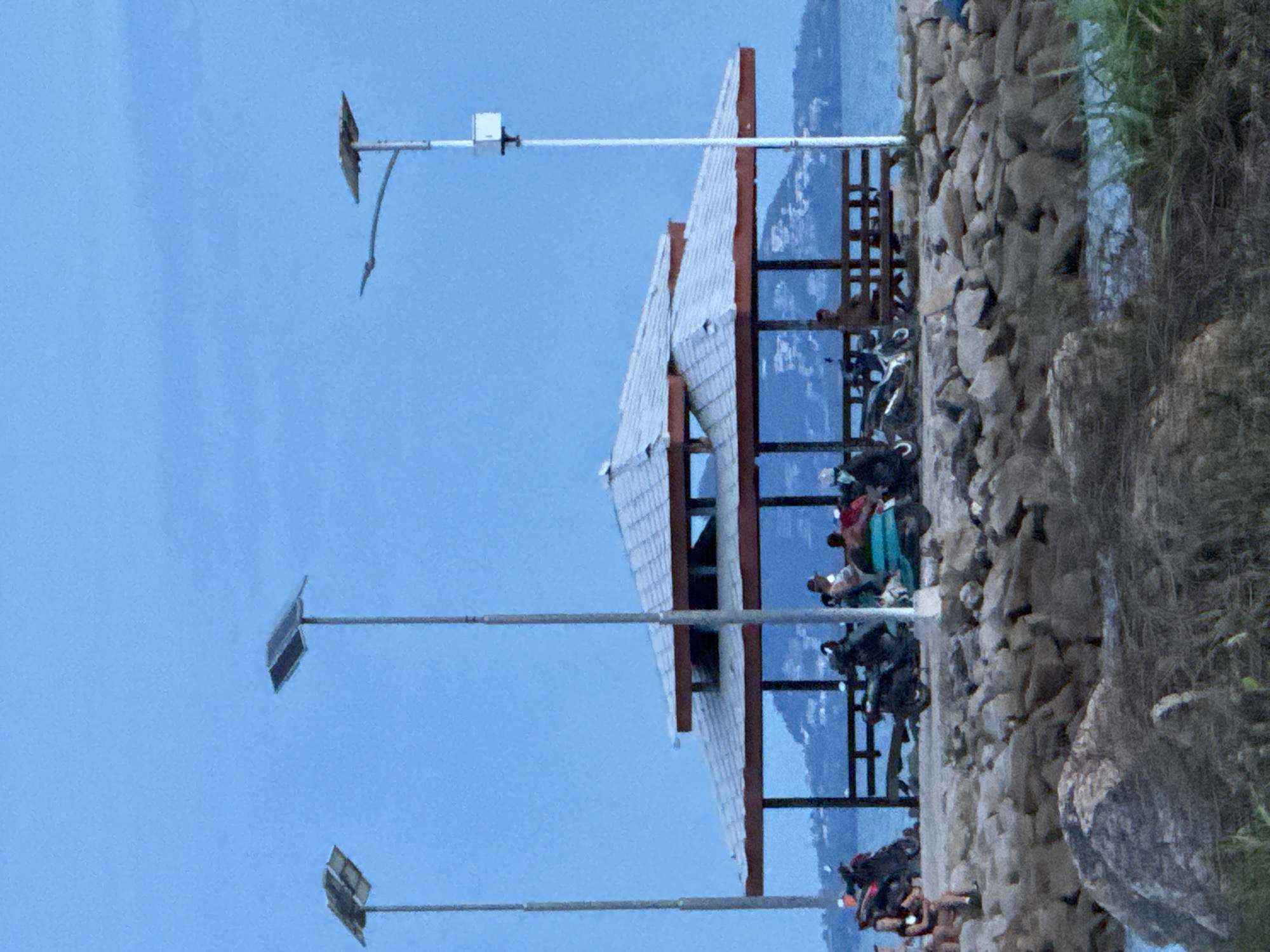 A photograph shows a three-story structure at the edge of a rock barrier. The structure, believed to be in Baan Tai, Thailand, has a gray-tiled roof and a frame made of dark-stained wood. The structure appears to be a viewing area, with individuals resting and socializing. Three pole structures with gray solar panels atop them are also visible in the image. The background includes a body of water, a mountain range, and a clear, bright blue sky.