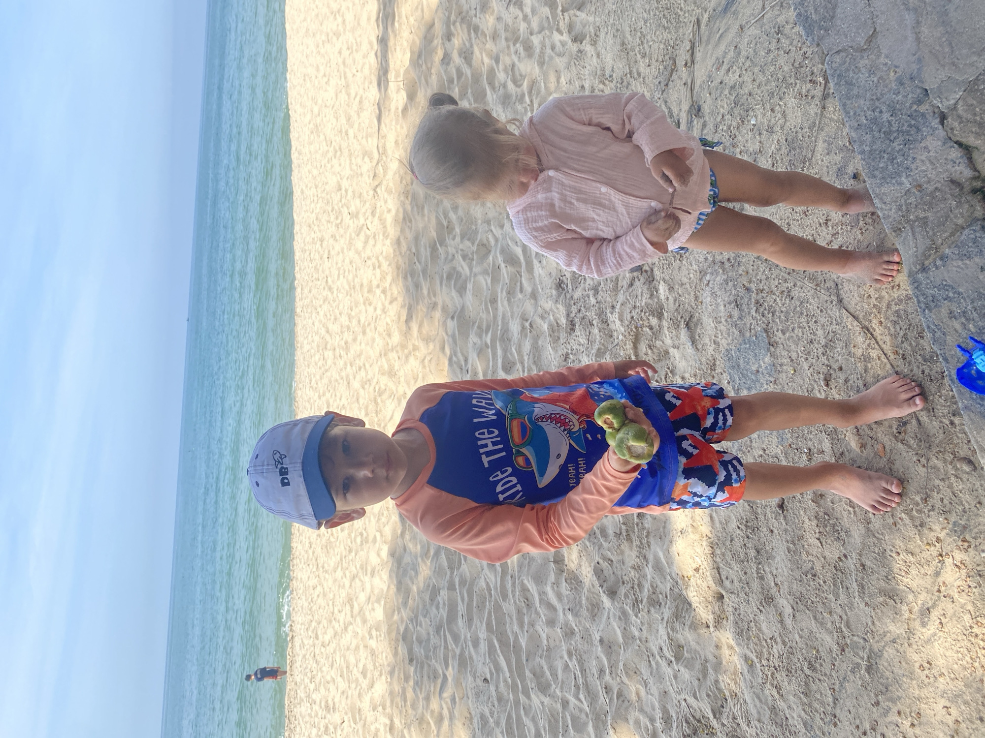 In Phu Thuy Ward, Vietnam, two children are standing on a sandy beach near the ocean. The boy in the foreground, wearing a blue and orange rash guard with a shark graphic and patterned swim trunks, holds a few green fruits in his hand. He is also wearing a blue and white baseball cap. To his right, a younger girl in a light pink cardigan and striped swimsuit looks down at her hands, holding a twig. The ocean stretches out behind them, with a calm blue surface meeting the horizon under a clear sky. The sand is light-colored and appears dry, with some shadows cast from unseen structures. In the distance, another person is visible standing in the shallow water.