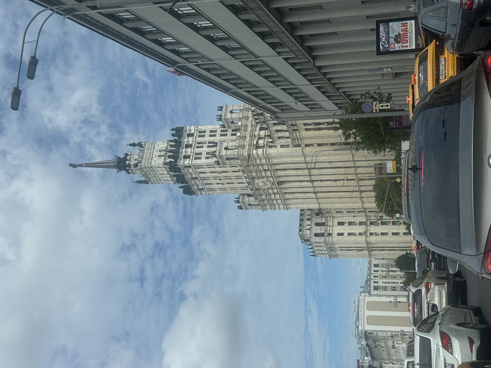 The image captures a vertical view of a city street in Moscow, Russia, with a focus on architectural grandeur and urban traffic. The foreground is dominated by cars in heavy traffic, suggesting a busy time of day. Among the vehicles, a yellow taxi is visible.
The middle ground features a strikingly ornate, light-colored building with numerous windows and intricate decorative elements, characteristic of Stalinist architecture. This building appears to be a prominent landmark, possibly a ministry building or a residential skyscraper. Its imposing presence is accentuated by the sky above, which is a mix of blue and white clouds, indicating fair weather.
Further in the background, another large building with a more modern, grey facade stretches along the street. Overhead street lamps with a distinctive design hang above the roadway. Visible text includes a billboard with Russian text, "Знать" and "ГОМАН", and a license plate "A846CB197". The overall impression is one of a bustling urban environment with a blend of historical and contemporary architecture. The image seems to be taken from within a vehicle, looking upwards and sideways.
