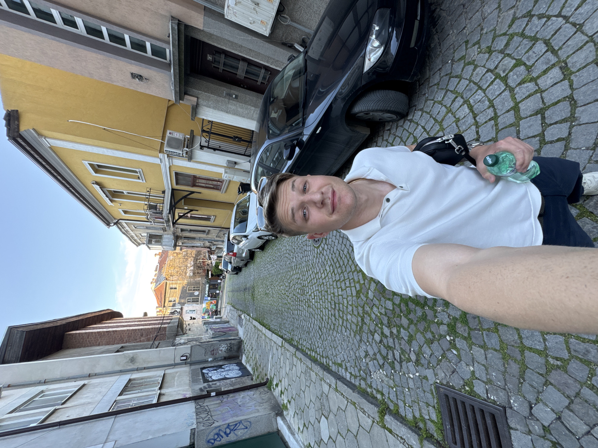A medium shot shows a fair-skinned man taking a selfie in Plovdiv, Bulgaria. He is holding a bottle of water in his right hand and wearing a white polo shirt. A small black bag is around his waist. The cobblestone street is narrow with cars parked alongside buildings of varying colors. The buildings are two to four stories tall. There is graffiti on some of the lower buildings. A street grate is visible in the foreground of the cobblestone street.