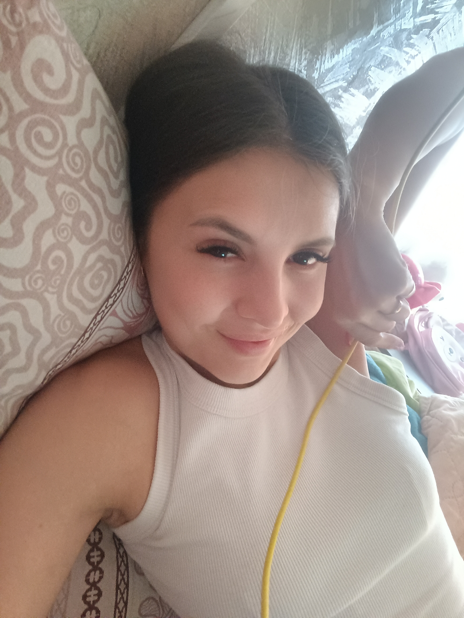 A young woman with dark hair pulled back from her face, wearing a white ribbed tank top, lies on a patterned pillow and bedding. A yellow cable is draped across her chest. The location is Unknown, Unknown.