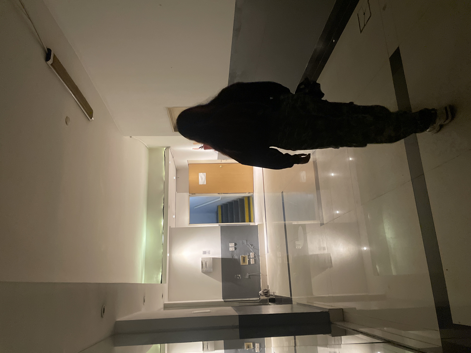 The image is taken in a hallway with a person walking from left to right. The hallway is brightly lit with white walls. Further down the hallway is a door leading to a stairwell. The person is wearing dark clothing. The floor is white tile. The location is not provided.