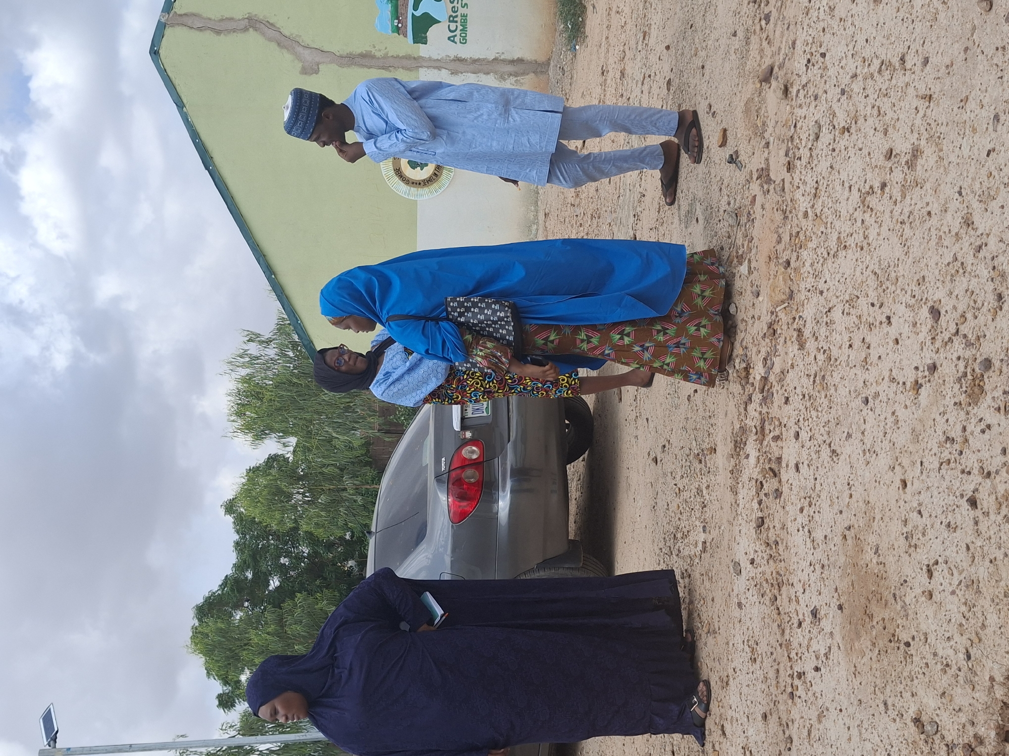 In Kundulun, Nigeria, under a cloudy sky, a group of four individuals stands near a light green building. On the left, a woman in a dark blue hijab and long dress stands with her back to the viewer, holding a notebook. To her right, two women in hijabs are conversing. One wears a blue hijab and a patterned dress, carrying a black handbag. The other, partially obscured, wears a blue garment and glasses. On the far right, a man in a light blue traditional outfit and a patterned cap stands, looking to his left with his hand on his chin. A grey Toyota car is parked behind the women, with its rear visible. The ground is a mix of sand and small stones. A street light with a solar panel is visible in the background. The building has a green trim along the roofline. A sign on the building reads "ACRes GOMBE ST".