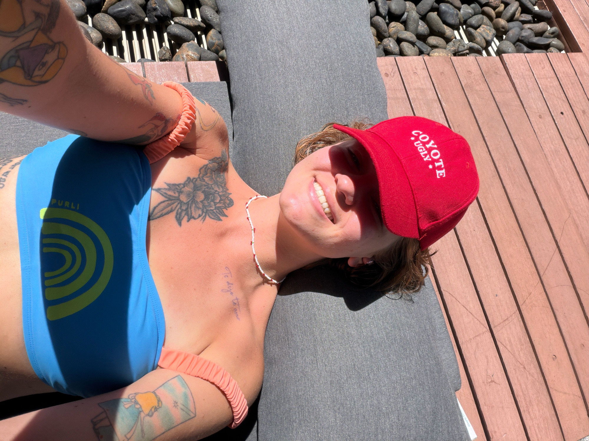 A person wearing a blue bikini top with a rainbow design and a red "Coyote Ugly" baseball cap lies on a gray lounge chair next to a wooden deck and a pile of black stones. The person has tattoos on their arms and chest and is wearing a white and red beaded necklace. Sunlight casts shadows on their face and body. The scene appears to be outdoors in a sunny environment. There are no discernable elements that place the location specifically within Hua Hin City Municipality, Thailand.