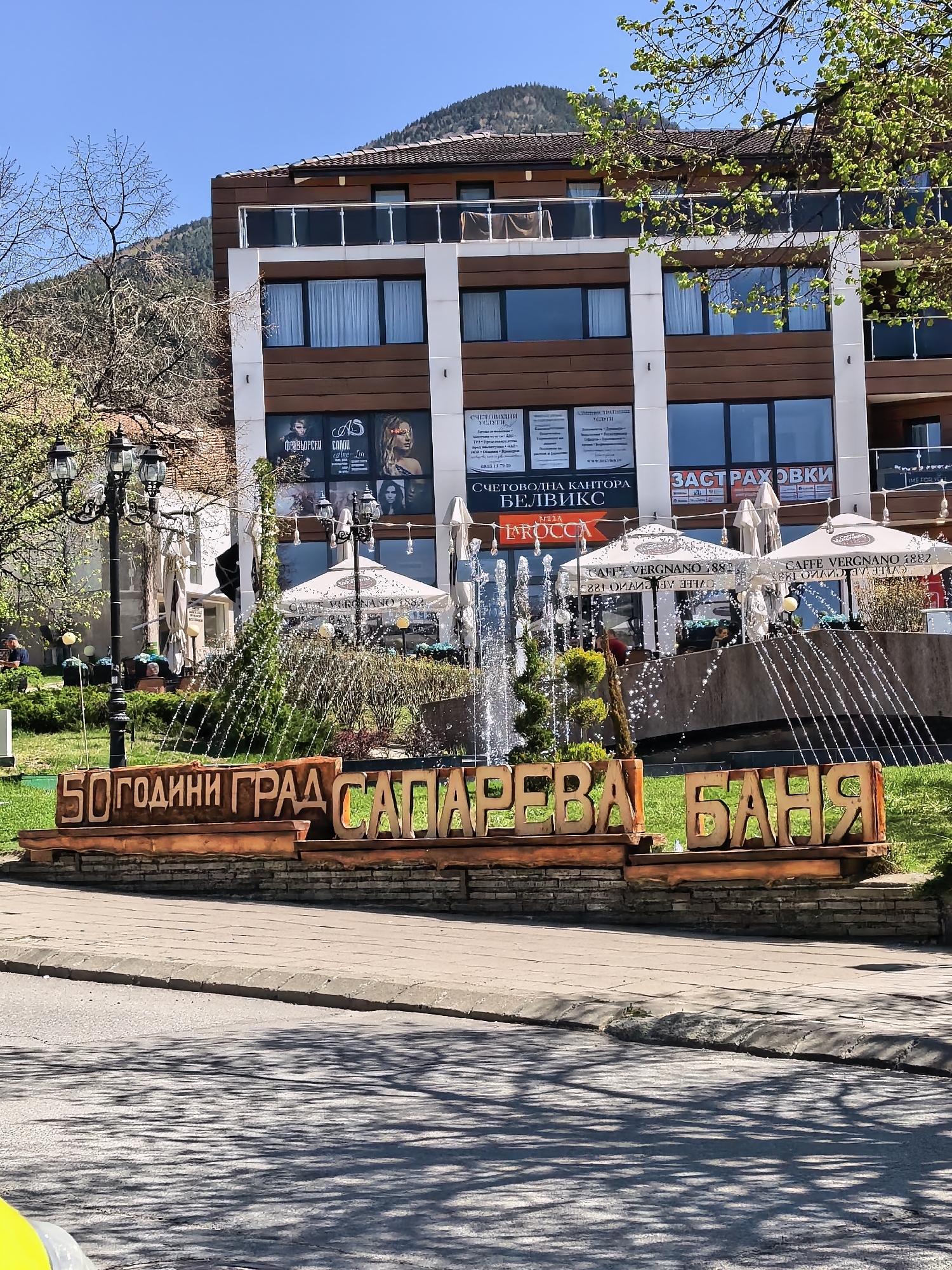 The image depicts a scenic view in Sapareva Banya, Bulgaria, featuring a sign that reads "50 години град Сапарева баня" (50 Years City Sapareva Banya), a fountain, and a building behind. The sign is made of wooden letters and is situated on a low stone wall bordering a grassy area. A fountain with several streams of water is located behind the sign, creating a shimmering effect.
Behind the fountain, there is a modern building with a combination of dark wood and white facade. There are business advertisements on the building's ground floor, under windows. Tables and umbrellas from Caffe Vergnano 1882 are visible on a raised terrace behind the fountain. A street lamp is situated to the left.
The sky is clear and blue, suggesting it is daytime. The surrounding trees have fresh spring leaves, indicating a pleasant climate. The foreground shows a paved road.