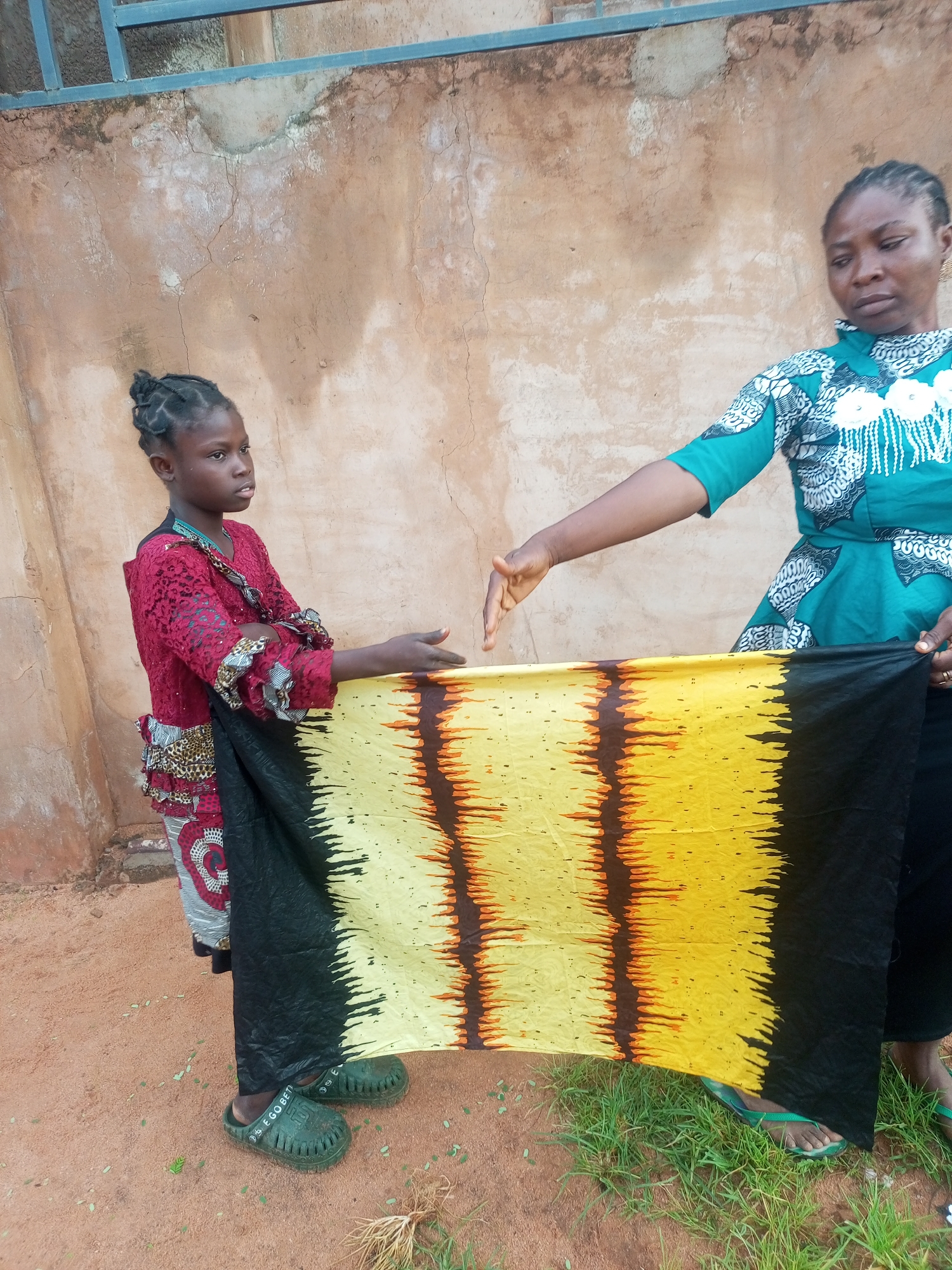 In Tempure, Nigeria, a young girl and an adult woman stand outside a building. The girl, wearing a red lace top and a patterned skirt, holds a black, yellow, and brown striped fabric. The adult woman, in a teal and white patterned dress, gestures towards the fabric with her right hand. Both are standing on reddish-brown dirt and scattered green grass, with a textured, beige-colored wall behind them. A blue railing is visible at the top of the frame.