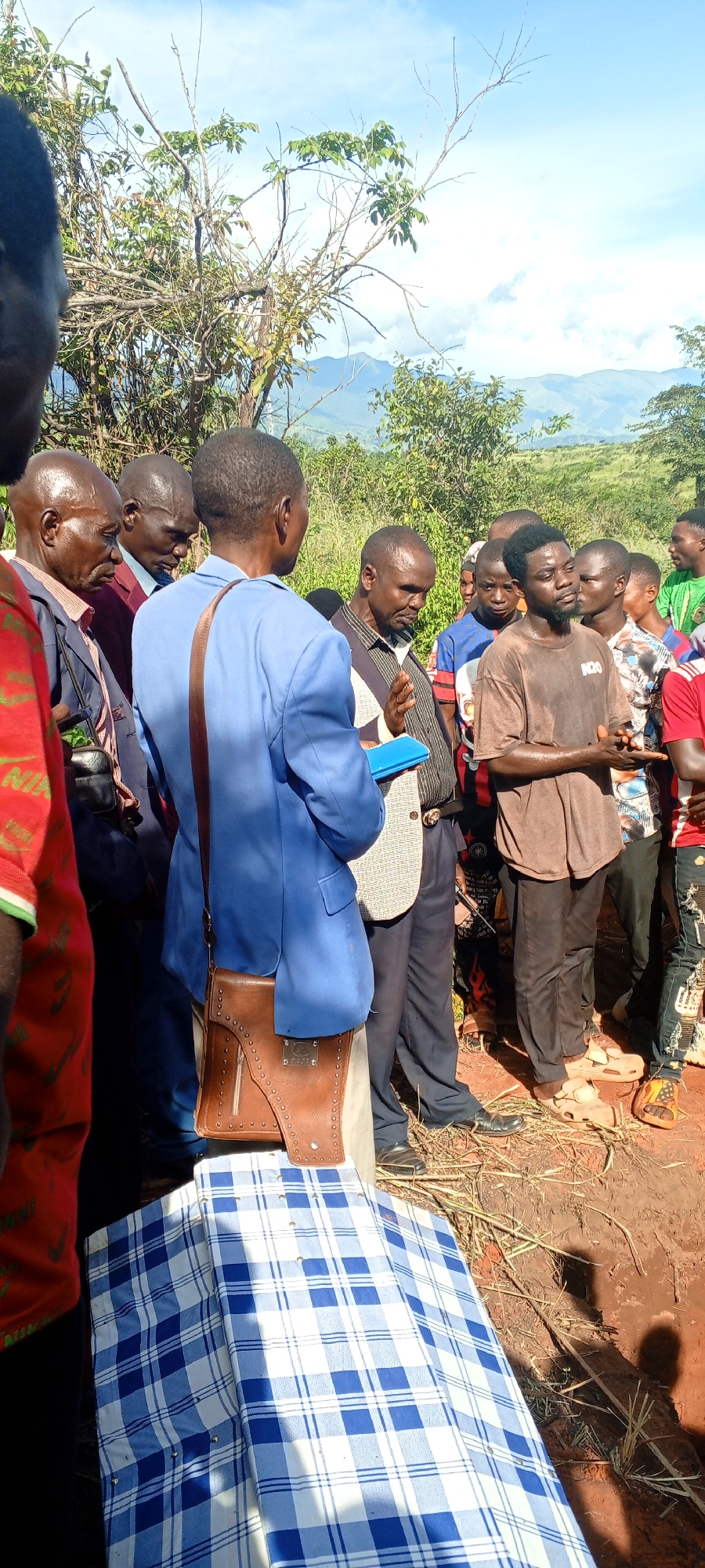 A group of people are gathered outdoors, near what appears to be an open grave. In the foreground, a coffin is covered with a blue and white checkered cloth. The group primarily consists of adult men of African descent, dressed in a variety of clothing from formal attire to casual wear. One man in a blue suit holds a blue document or book. The environment appears rural, with trees, brush, and a mountain visible in the background. The scene suggests a funeral or memorial service. The location is consistent with being in the vicinity of Ngalula, Democratic Republic of the Congo, based on the presence of indigenous Africans and a rural environment.