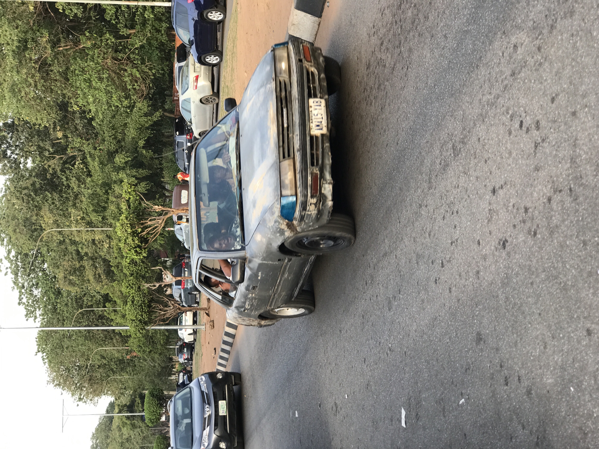 The image depicts a busy road scene in Abuja, Nigeria, during what appears to be late afternoon or early evening. The prominent subject in the midground is an older, grey sedan showing signs of wear and minor body damage. Its front license plate, "AY 15-YAB," is clearly visible. Two occupants, including the driver and a rear passenger, are partially visible inside the car.
In front of the grey sedan, a modern black SUV, possibly a Toyota RAV4, is partially seen. The road surface is dark asphalt, and a black and white striped curb or barrier lines the right side of the road.
The background features a line of diverse vehicles, including blue, white, and silver cars, suggesting either parked cars or a traffic queue. The street is flanked by dense, tall green trees, indicating a well-vegetated environment. Several modern street light poles are also visible extending along the road. The overall weather appears clear and dry, and the natural lighting creates a soft, warm ambiance without harsh shadows. The scene captures typical urban traffic and infrastructure in the Nigerian capital.