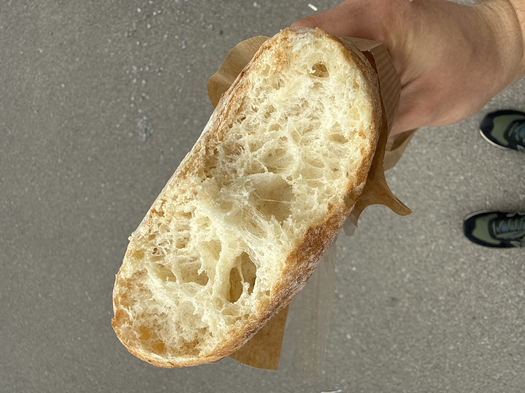 A person is holding a large slice of bread in a brown paper bag. The bread has a golden-brown crust and a light-colored, airy interior. The person is holding the bread above a gray concrete surface with speckled discoloration. The tips of a pair of running shoes in muted earth tones are partially visible in the lower right corner of the image. The location of the photo is unknown, and cannot be located in the city of Sofia, Bulgaria based on the image alone.