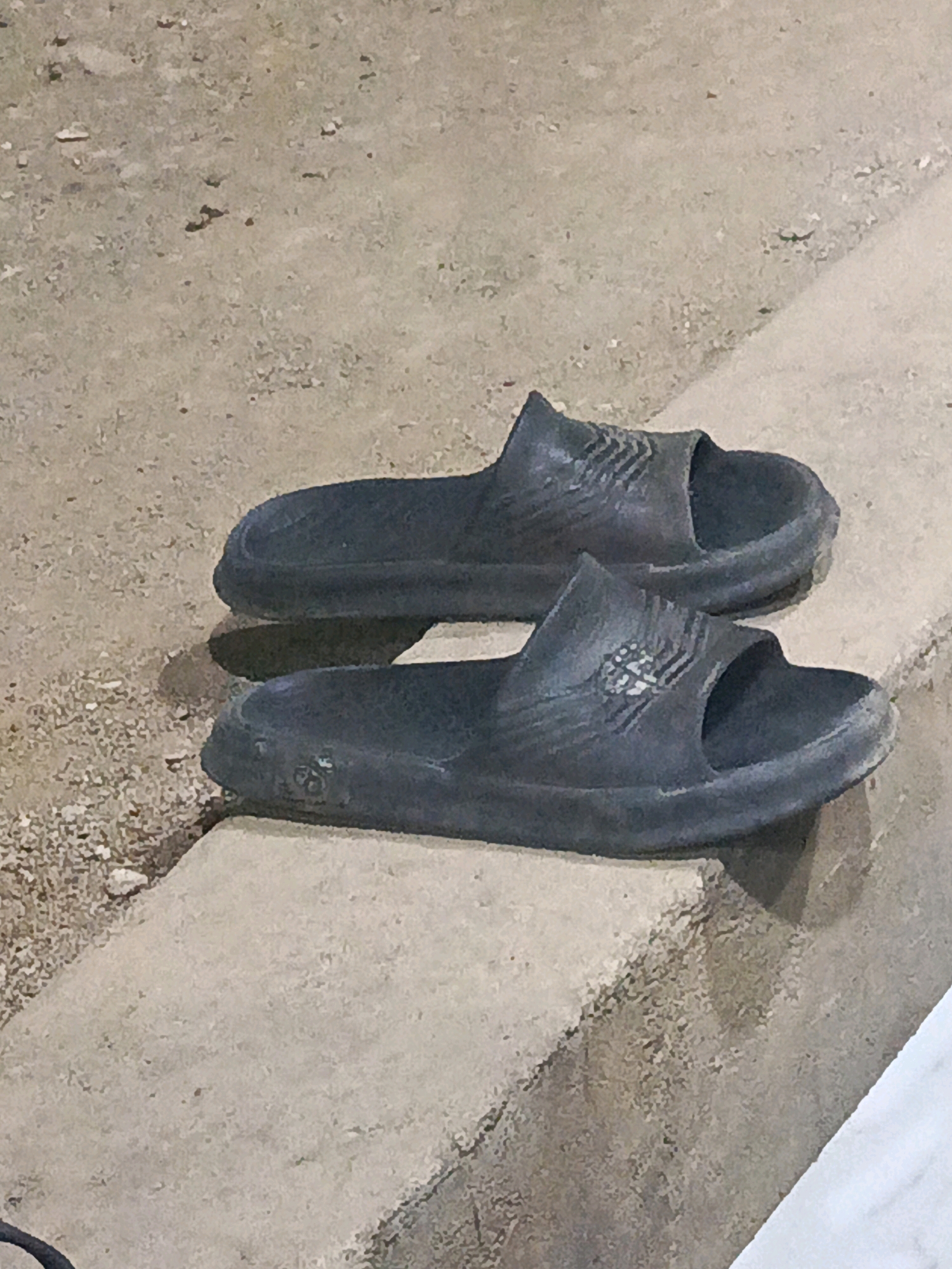 Two black slide sandals are positioned side-by-side on a concrete ledge. The sandals are facing forward, with the soles down and the straps visible. The ground behind the ledge is a textured, sandy surface with some scattered debris. The lighting suggests it might be dusk or an indoor setting with artificial light. The scene is devoid of human presence. This image was captured in Megang Sakti V, Indonesia.
