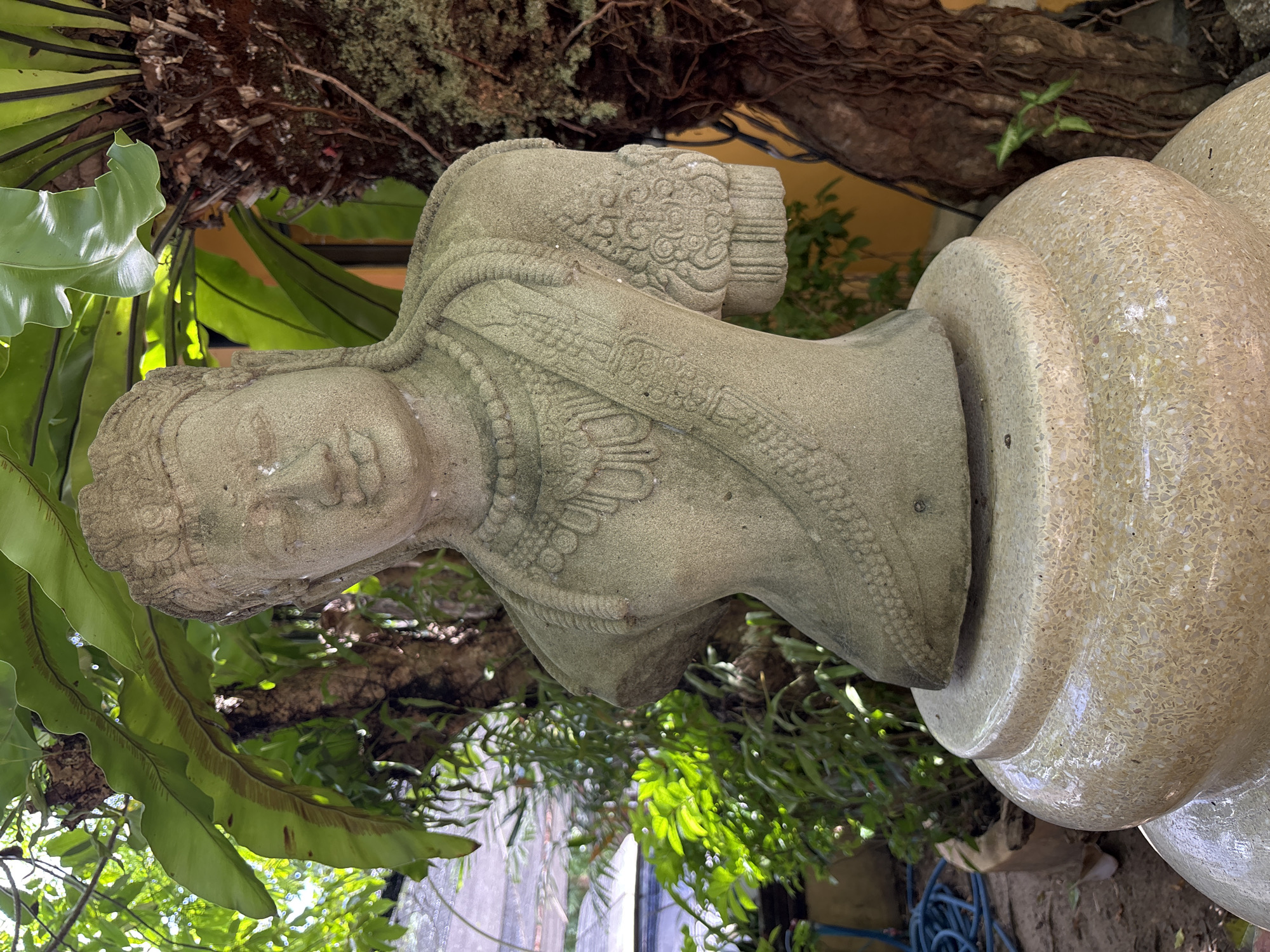 The image shows a stone statue of a bust in a garden setting. The statue depicts a figure with elaborate headdress and jewelry, including a beaded necklace and armbands. The figure's arms are missing below the elbows. The statue is placed on a multi-tiered pedestal. Behind the statue are lush green tropical plants, including large fern leaves and the trunk of a tree with exposed roots. The background is blurred, but hints of a yellow wall and a white canopy are visible. A blue garden hose is visible in the lower left corner. The lighting suggests it is daytime. The image appears to have been taken in Chalong, Thailand, likely in a temple or a private garden.