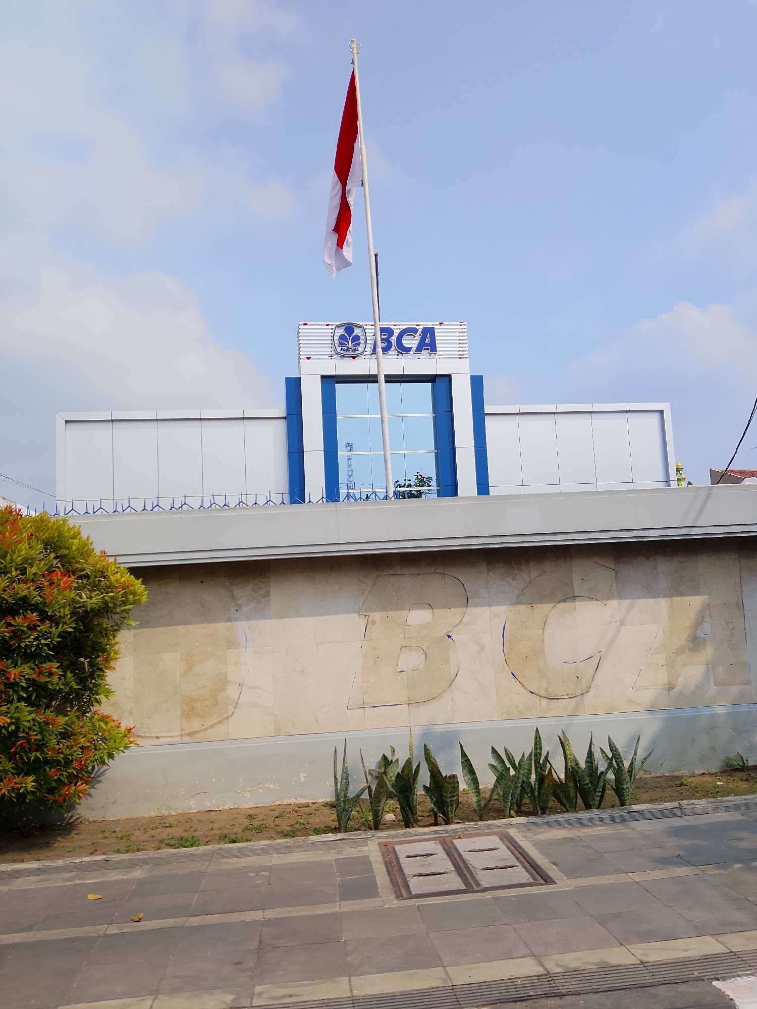 A low-angle, eye-level, outdoor, daylight, medium shot captures the facade of a building in Balun, Indonesia. The building features a modern architectural style, characterized by clean lines and a white and blue color scheme. A prominent Indonesian flag, red and white, flies from a tall flagpole mounted on top of the building. The building's signage displays the blue logo and text "BCA," indicating it is likely a Bank Central Asia branch. Below the signage, a large glass-paneled window reflects the sky and surrounding elements. The building is set against a backdrop of a bright blue sky with scattered white clouds.
In the foreground, a section of a paved walkway is visible, with a line of snake plants bordering a low wall. The wall itself is adorned with large, light-colored stone tiles and features embossed or painted large letters, partially visible, that appear to spell out "BCA" again, possibly an older or secondary sign. To the left, a lush green bush with hints of red foliage adds a touch of nature to the urban landscape. The overall scene is clean and well-maintained, suggesting a public or commercial space. No people are visible in the frame.