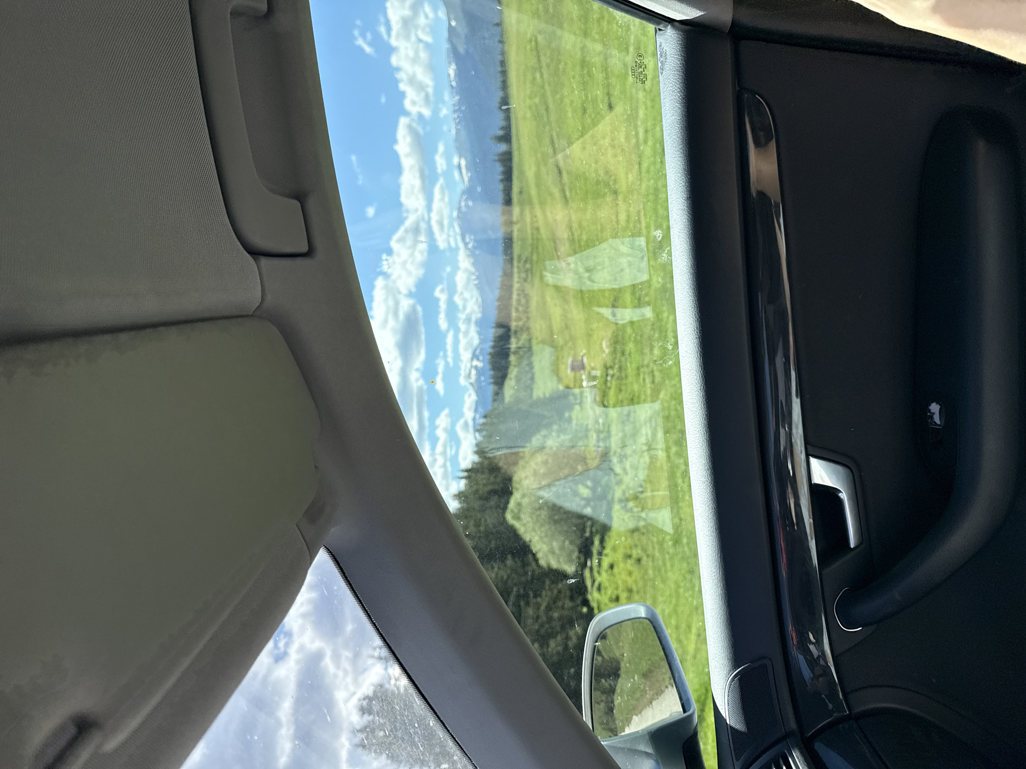 The image is taken from inside a car, looking out the passenger window and windshield. The interior of the car is visible in the foreground, including a portion of the door panel with a black leather finish, a chrome-colored door handle, and a speaker grille. Above the window, the car's headliner and part of the sun visor are visible.
Outside the car, a bright, sunny day is depicted. The view is of a lush green meadow with rolling hills. In the mid-ground, a dense forest of dark green trees is visible, likely pine or fir. Beyond the forest, a range of majestic, snow-capped mountains stretches across the horizon under a bright blue sky dotted with fluffy white clouds. There are reflections on the car window, suggesting the presence of a building or structure within the meadow, possibly a house or shed. The side mirror of the car is also visible, reflecting a dirt path and more green landscape.
The image does not provide any specific information or visual cues that would definitively place it in the city of Avramovo, Bulgaria.