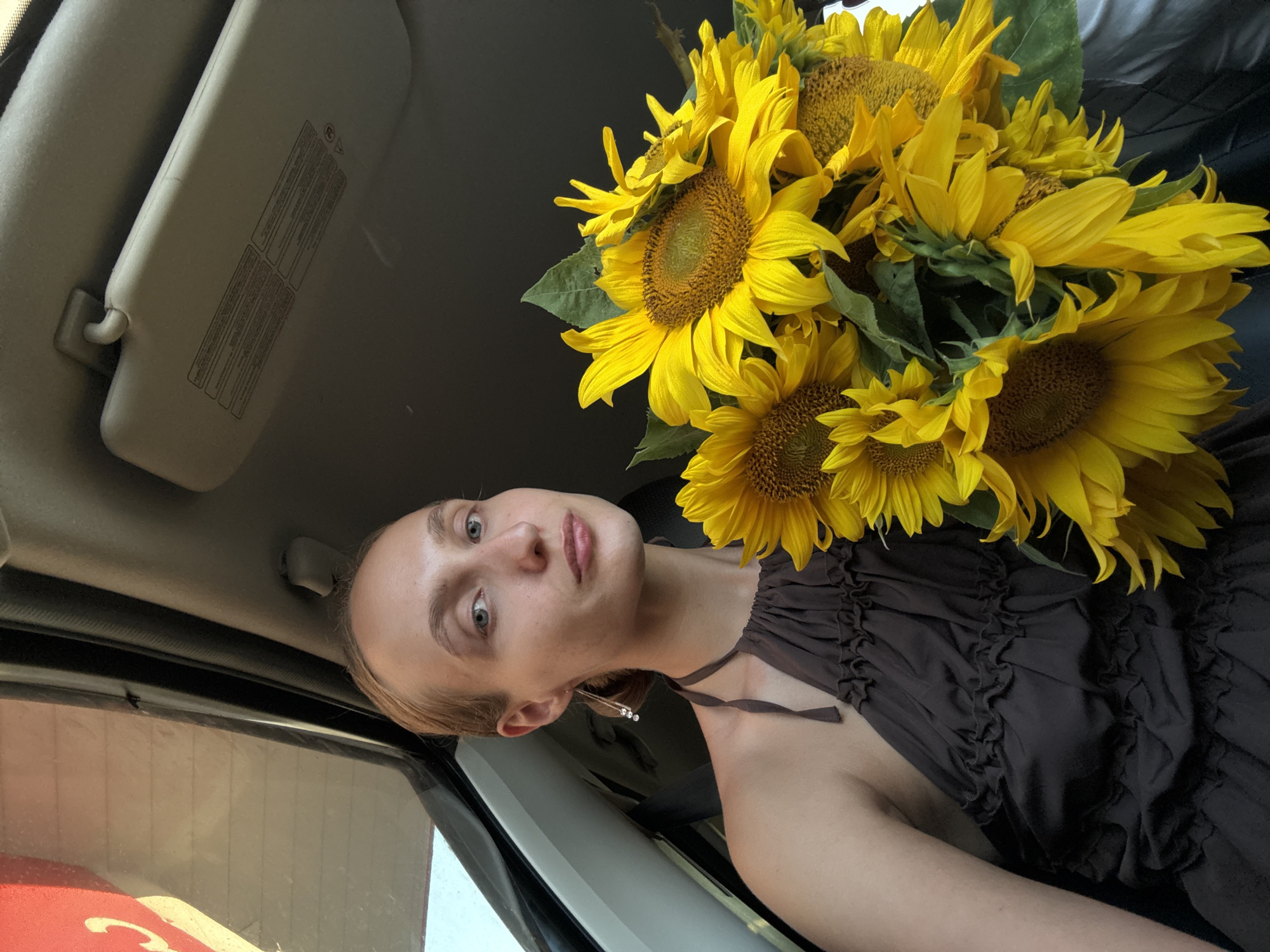 A young person with light skin and blue eyes is in the passenger seat of a car, holding a large bouquet of sunflowers. The person is wearing a dark brown dress with ruffled detailing and thin straps. They have a delicate earring with three small gems dangling from their ear. The person's expression is neutral, with a slight upward gaze towards the camera.
Sunlight is visible through the car window, suggesting it is daytime. The interior of the car is visible, including a sun visor with text printed on it. The text on the sun visor is small and illegible in the provided image, but appears to be informational or warning labels. The sunflowers are vibrant yellow with brown centers, and some appear to be wilting slightly.
The scene is a close-up shot, with the person and the sunflowers filling most of the frame. The background is dark, likely indicating the interior of the car or the surrounding environment being out of focus. The overall mood is calm and natural, with the bright sunflowers providing a cheerful contrast to the darker elements of the scene. The image appears to have been taken in a natural light setting, likely during the day. No specific weather conditions are discernible.