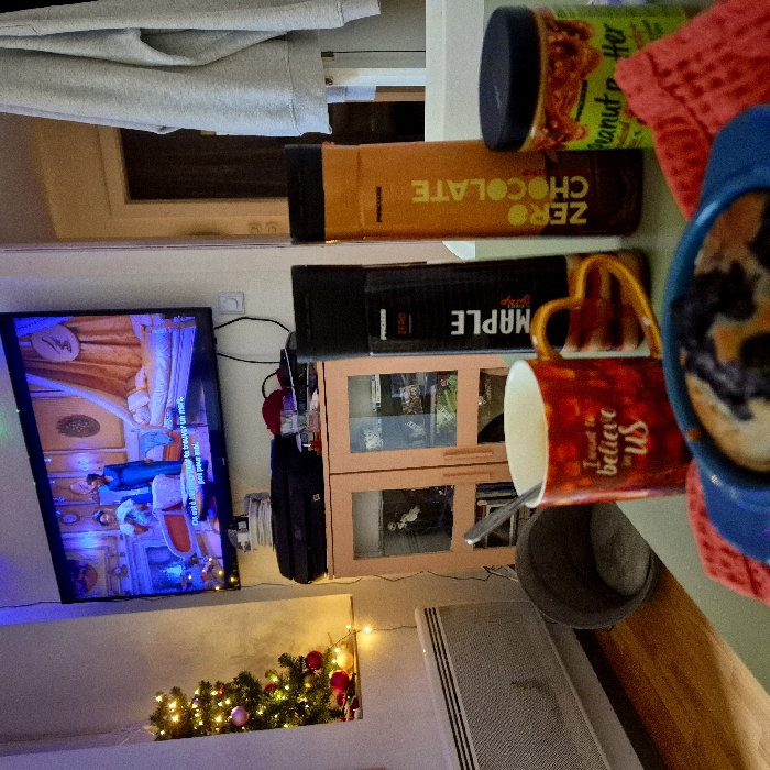 This image depicts an indoor domestic setting, likely a living room or studio apartment. The specific location within Strasbourg, France, is not identifiable from the visual evidence.
In the foreground, a light green table surface holds several items: A blue ceramic bowl contains a baked good, possibly a crumble or muffin, with visible dark berries. Beneath the bowl is a textured red cloth or mat. To the left, a red ceramic mug with "I want to believe in US" text and a heart-shaped handle contains a light-colored beverage and a silver spoon. Behind the mug, a tall black bottle labeled "PROZIS ZERO MAPLE" stands next to a tall orange bottle labeled "PROZIS ZERO CHOCOLATE." A jar, partially obscured, with a green label and "Peanut Butter" text is also on the table.
The midground features a wall-mounted flat-screen television displaying a scene with two women in period clothing within an opulent room; French subtitles are visible, reading "On est là à trouver un mot, pour moi." Below the TV, a two-door wooden cabinet with glass-fronted upper sections holds various items. A black printer rests on top of this cabinet, alongside a red cap and a small glass. To the left of the cabinet, a small, illuminated Christmas tree with warm white lights and red ornaments stands near a window. Below the window, a white horizontal radiator is visible. On the wooden floor, a grey cylindrical pet bed or basket is situated. String lights with warm white bulbs are strung along the wall below the Christmas tree.
In the background to the right, a grey hooded sweatshirt or jacket hangs on a white rack, partially visible within a doorway or opening. The room is illuminated by ambient light, the television screen, and decorative string lights and Christmas tree lights, with a subtle blue hue from an overhead light source. No people are directly visible within the room, only characters on the television screen.