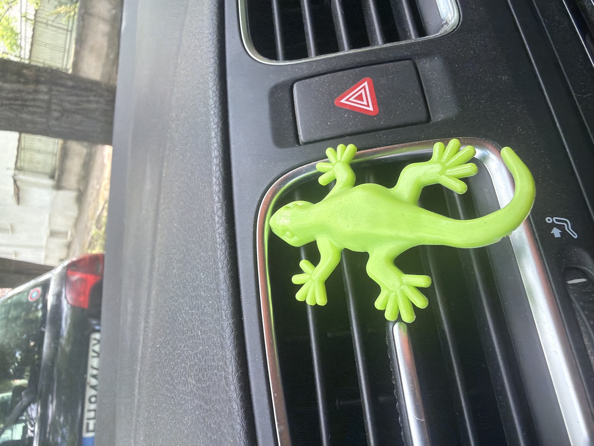 The image displays the interior of a car, specifically a close-up of the dashboard and an air vent, located in Iskar, Bulgaria. A prominent bright neon green, gecko-shaped object, likely an air freshener or decoration, is attached to the chrome-trimmed air vent. The object appears to be made of soft plastic or rubber, featuring distinct toes and a curled tail. Above the vent, a black button for the hazard warning lights is visible, marked with a red triangle symbol. The dashboard material is a dark, textured grey or black.
Through the car window, an outdoor scene is observed, suggesting the car is parked or stationary during daylight hours. Trees with green leaves are reflected or seen in the background, indicating a sunny day. On the left side of the frame, the rear of another dark-colored vehicle is partially visible, showing its red taillight and a distorted view of its license plate. Visible Cyrillic characters on the license plate, such as "Л", "О", and "А", are consistent with a Bulgarian registration format, confirming the provided location context. No people are visible, and no specific activity is taking place other than the static display of the car's interior.