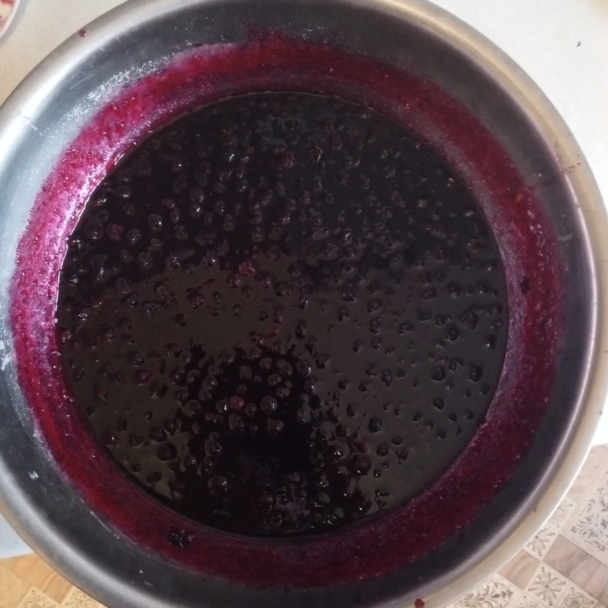 The image shows a close-up, overhead view of a stainless steel bowl filled with dark, viscous liquid and small, round, dark berries, likely blueberries or similar. The liquid has a deep purple hue and appears to be a compote or jam. The edge of the bowl has a thick, darker purple residue, possibly from the cooking process. The bowl is placed on a surface that appears to be a tiled floor with a patterned design. The lighting is overhead, casting soft shadows. There are no people or animals visible in the image, nor any specific identifying landmarks that would place it in Sayanogorsk, Russia.