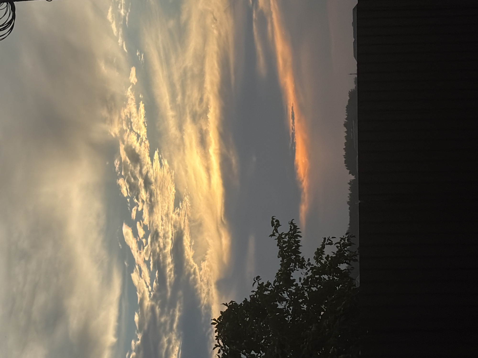 The image depicts a sunset scene in Vysokaya Gora, Russia. In the foreground, a dark, solid fence stretches across the bottom of the frame, casting a shadow over the lower portion of the image. To the left, a silhouetted tree with lush green foliage extends into the midground. The background features a distant treeline, a hint of what appears to be a roofline, and a pole. The sky is the dominant element, filled with dramatic clouds illuminated by the setting sun. Wispy, light-colored clouds are interspersed with darker, heavier clouds, creating a textured sky with varying shades of grey and yellow. A bright orange glow emanates from a band of clouds near the horizon, suggesting the direct rays of the setting sun. The overall impression is one of a tranquil, late evening in a suburban or rural setting.