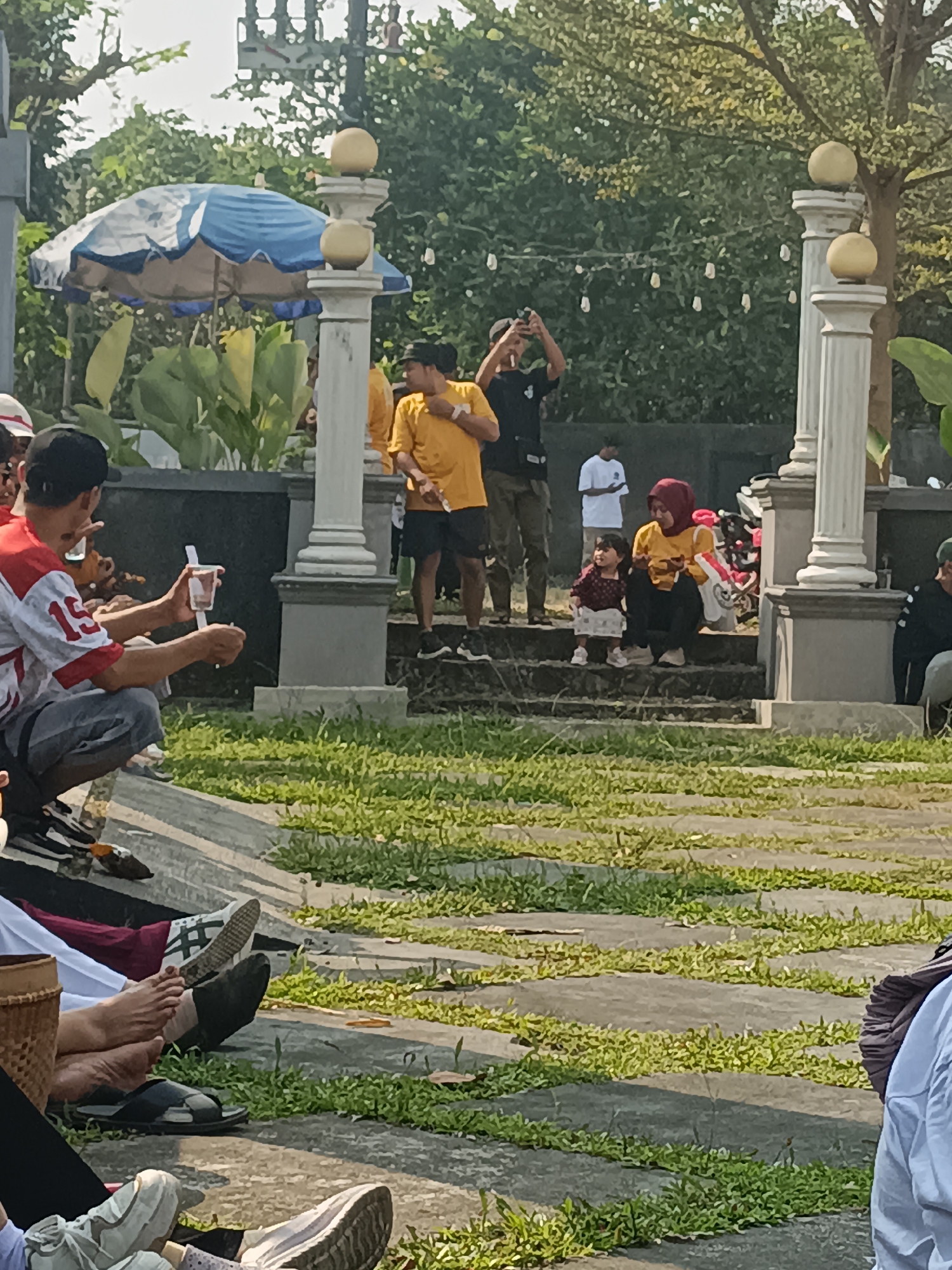 In a sunlit outdoor area in Sleman, Indonesia, a gathering of people are observed. In the foreground, several individuals are seated or crouching on a pathway bordered by grass. One person in a red and white shirt, identified by the number "15" on their sleeve, is crouched and holding a clear plastic cup with a white straw. To the right, a woman wearing a red hijab and a yellow shirt sits on a set of concrete steps with a young child standing beside her. The child is wearing a patterned top and a light-colored skirt. Behind them, other individuals are standing, including one man in a black t-shirt holding a phone to take a picture. The environment features decorative pillars with golden spheres, lush greenery, and a blue and white umbrella. The overall scene suggests a casual outdoor event or gathering.