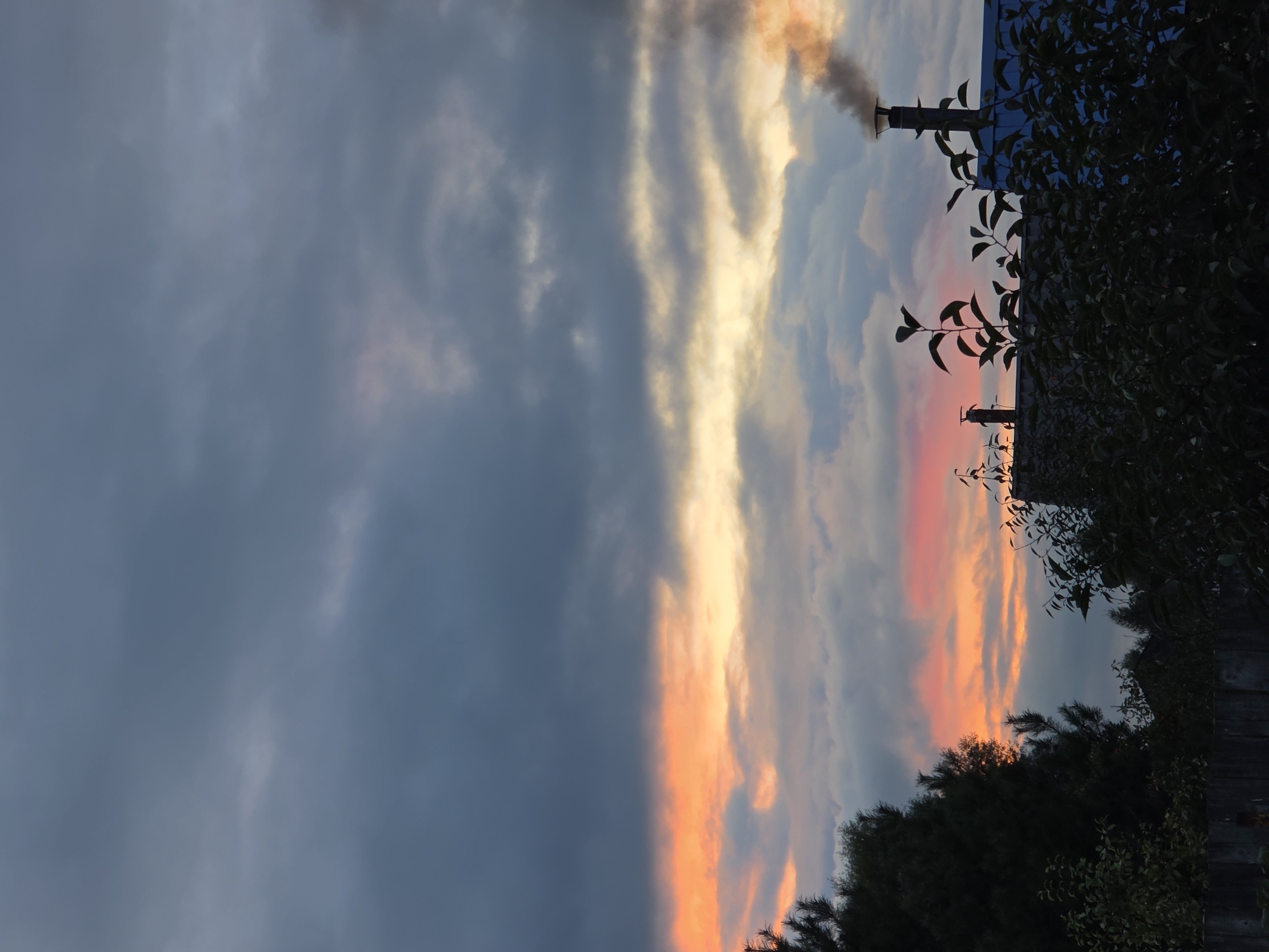 A photograph captures a dramatic sunset over a rural landscape, possibly in Речицы, Russia. The sky is a canvas of deep grays and blues, with a vibrant band of orange and pink hues illuminating the horizon. Wisps of clouds are scattered throughout, adding texture and depth to the celestial display. In the foreground, silhouetted against the colorful sky, are the dark shapes of trees and foliage. A fence runs along the bottom of the frame, partially obscuring the lower portion of the scene. A house with a blue roof is visible, with two chimneys. One chimney emits a plume of dark smoke, suggesting a fire is burning. The overall impression is one of a serene, yet somewhat ominous, evening.