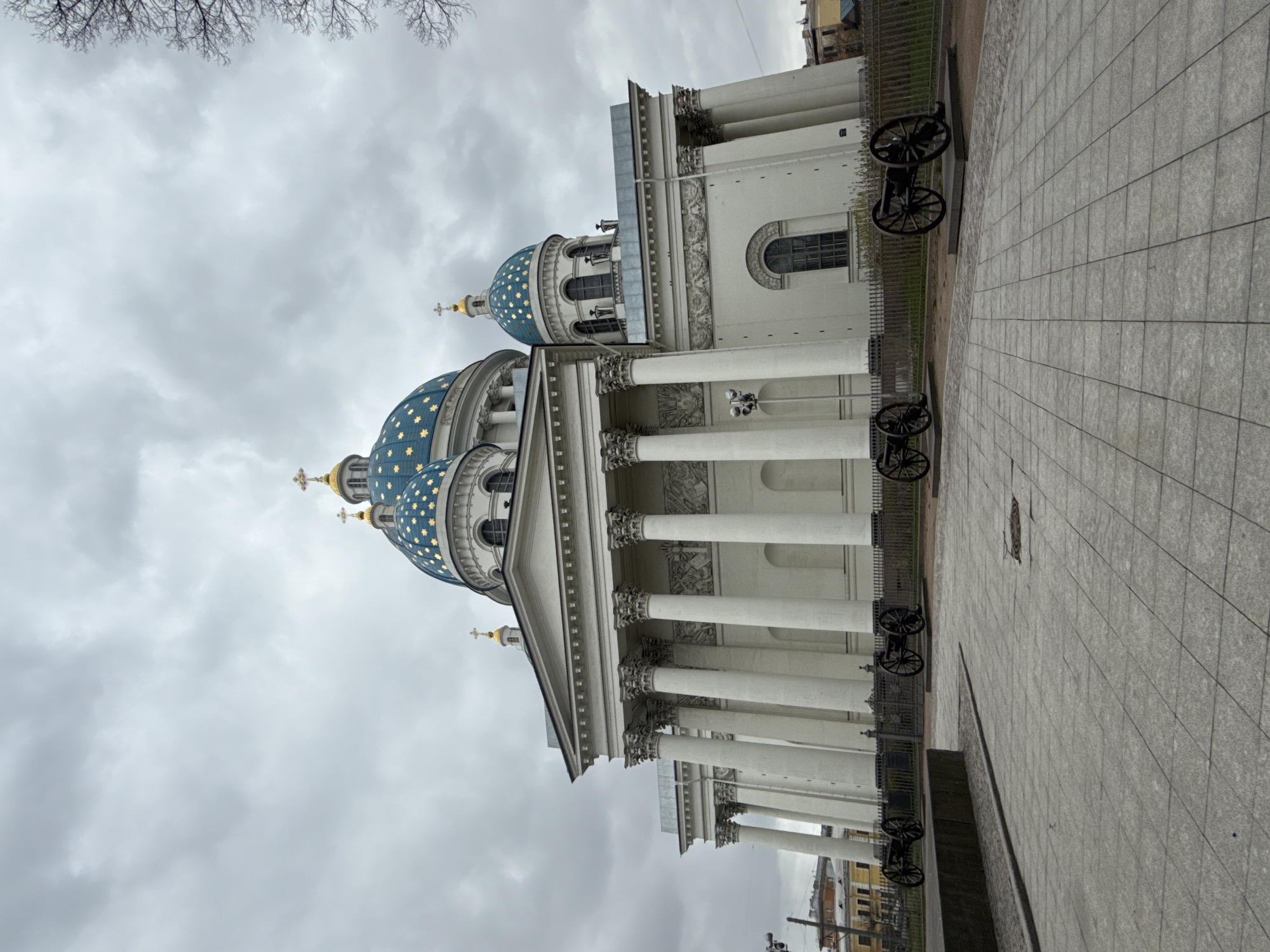 The image showcases the exterior of the Trinity Cathedral in Saint Petersburg, Russia. The cathedral, with its distinctive blue domes adorned with gold stars, is the focal point. The architecture features classical elements, including white columns and intricate detailing. The cathedral is surrounded by a paved area with cannons positioned along the fence. Above the cathedral, a cloudy sky dominates the scene. Bare tree branches are visible in the upper portion of the frame.