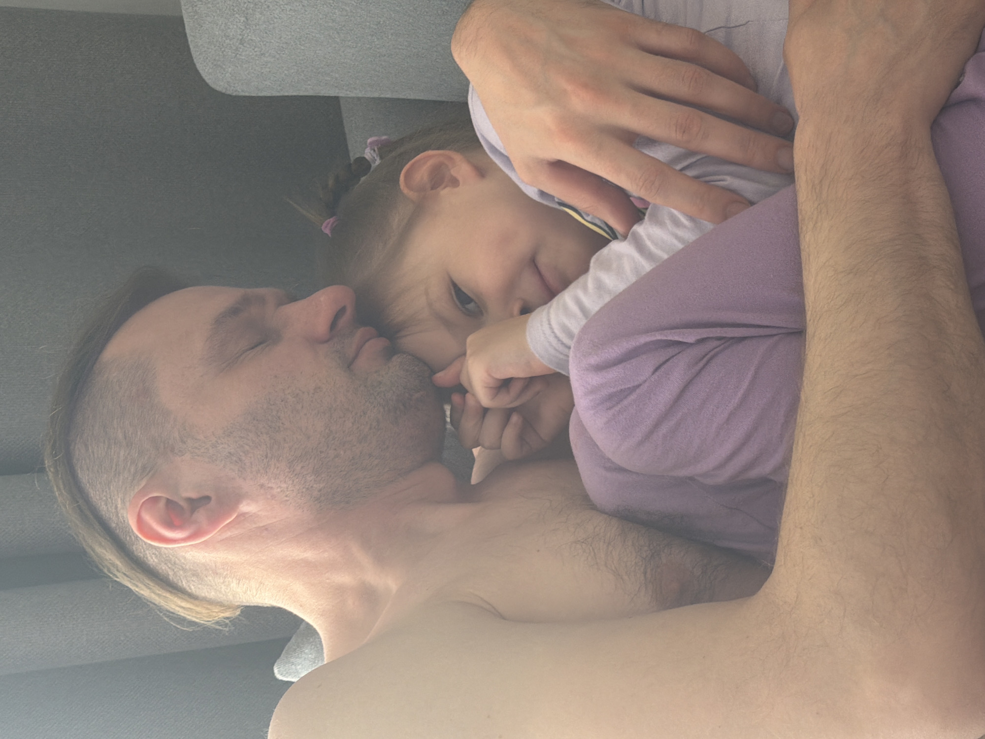 An intimate portrait captures a man and a young child cuddling closely on a grey couch or sofa. The image is oriented vertically, with the subjects positioned diagonally.
The man, appearing to be bare-chested with short facial stubble and longer hair on top, has his eyes closed, suggesting he is sleeping or resting peacefully. His left arm is wrapped around the child, holding her close. The child, likely a young girl, is snuggled against his chest and neck. She has light-colored hair styled in braids with pink hair ties visible. Her face is turned slightly towards the viewer, with one eye partially open, giving a soft, perhaps curious, gaze. She is wearing light-colored clothing, including a light purple top or pajamas and a lighter grey/white undershirt. Her hands are near the man's chest and neck.
The setting appears to be indoors, likely a home, with a grey couch or similar upholstered furniture item dominating the background. The lighting is soft and diffused, creating a gentle and warm atmosphere without harsh shadows. There are no direct indications of the time of day, but the relaxed postures and intimate closeness suggest a moment of rest or quiet bonding. No text is visible in the image. The overall mood conveys tenderness, comfort, and affection between the two individuals. The location context is Chaikovsky, Russia.