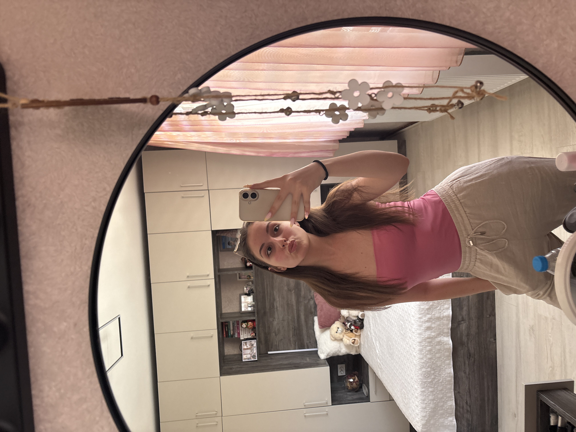 A young woman is taking a mirror selfie in a bedroom. She is wearing a pink strapless top and beige sweatpants. She has long brown hair and is puckering her lips. The room has a large bed with white bedding, a headboard with shelves displaying books and pictures, and a wall of built-in closets. A pink curtain is visible behind her. The mirror is round and framed in black. A hanging decoration with flower shapes is on the right side of the mirror. The floor is light-colored wood. The image appears to be taken indoors, likely in a residential setting in Bulgarovo, Bulgaria.