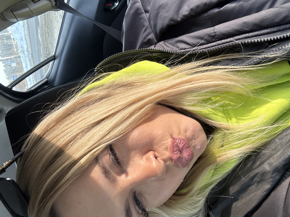 A close-up selfie taken inside a vehicle depicts a light-skinned female in L'vovskiy, Russia. The subject has long, straight blonde hair, with sunglasses perched atop her head. She is wearing a neon yellow hooded sweatshirt under a dark grey or black puffer jacket. Her eyes are partially closed, and her lips are pursed in a kissing gesture. Direct sunlight illuminates her face from the viewer's right. The interior of the car shows a dark seat and door panel. Through the car window, a winter environment is visible, featuring snow and ice on the ground, a road surface, and bare trees in the distance under a clear blue sky.
